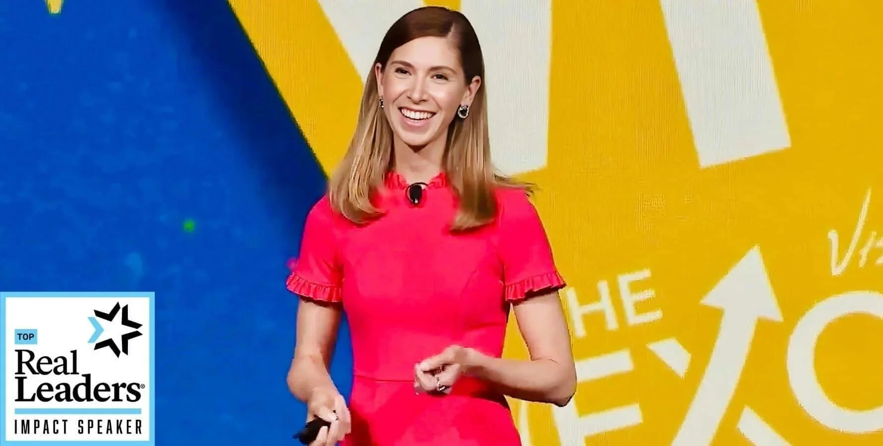Kristel Bauer with long brown hair smiling, wearing a bright pink dress with ruffled sleeves, standing on stage with a yellow and blue background featuring white and yellow graphics.