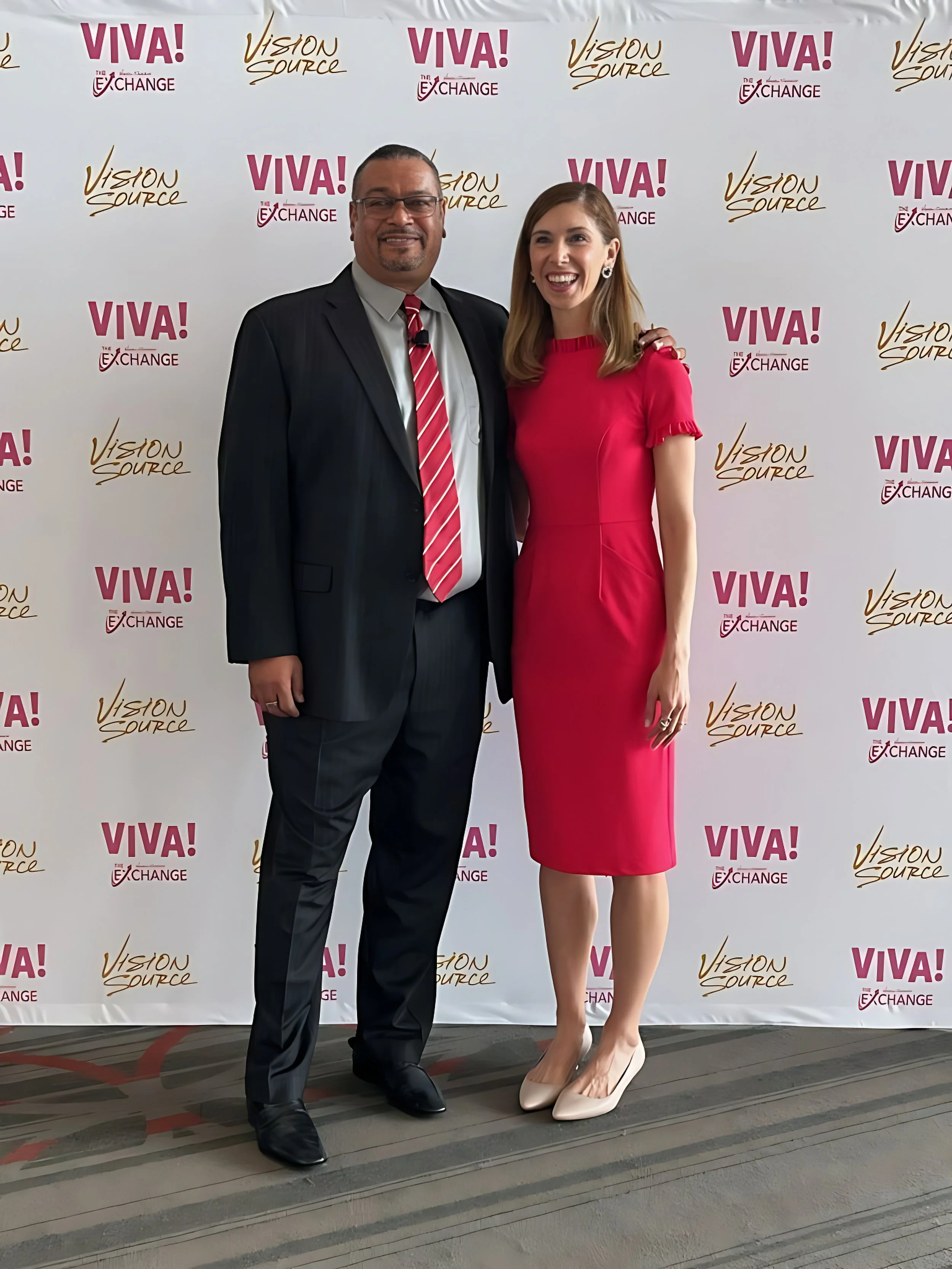 A man in a dark suit with a red and white striped tie and a woman in a pink dress stand together in front of a backdrop featuring logos that say "VIVA!", "The Exchange", and "Vision Source". They are smiling and appear to be at an event.