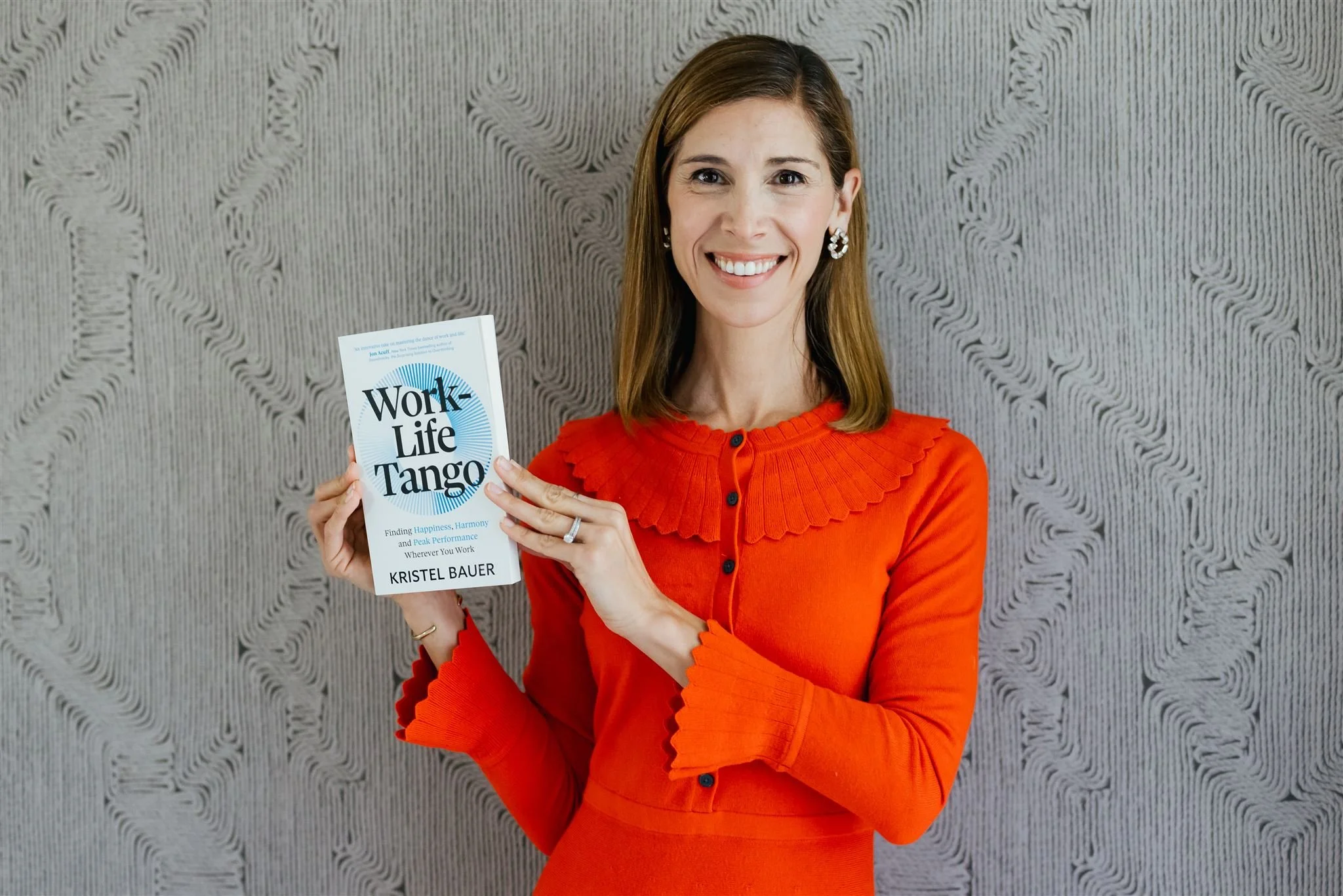 Kristel Bauer with shoulder-length brown hair, wearing a bright orange dress with ruffled collar and cuffs, holding a book titled "Work-Life Tango" by Kristel Bauer, standing in front of a textured gray wall and smiling at the camera.