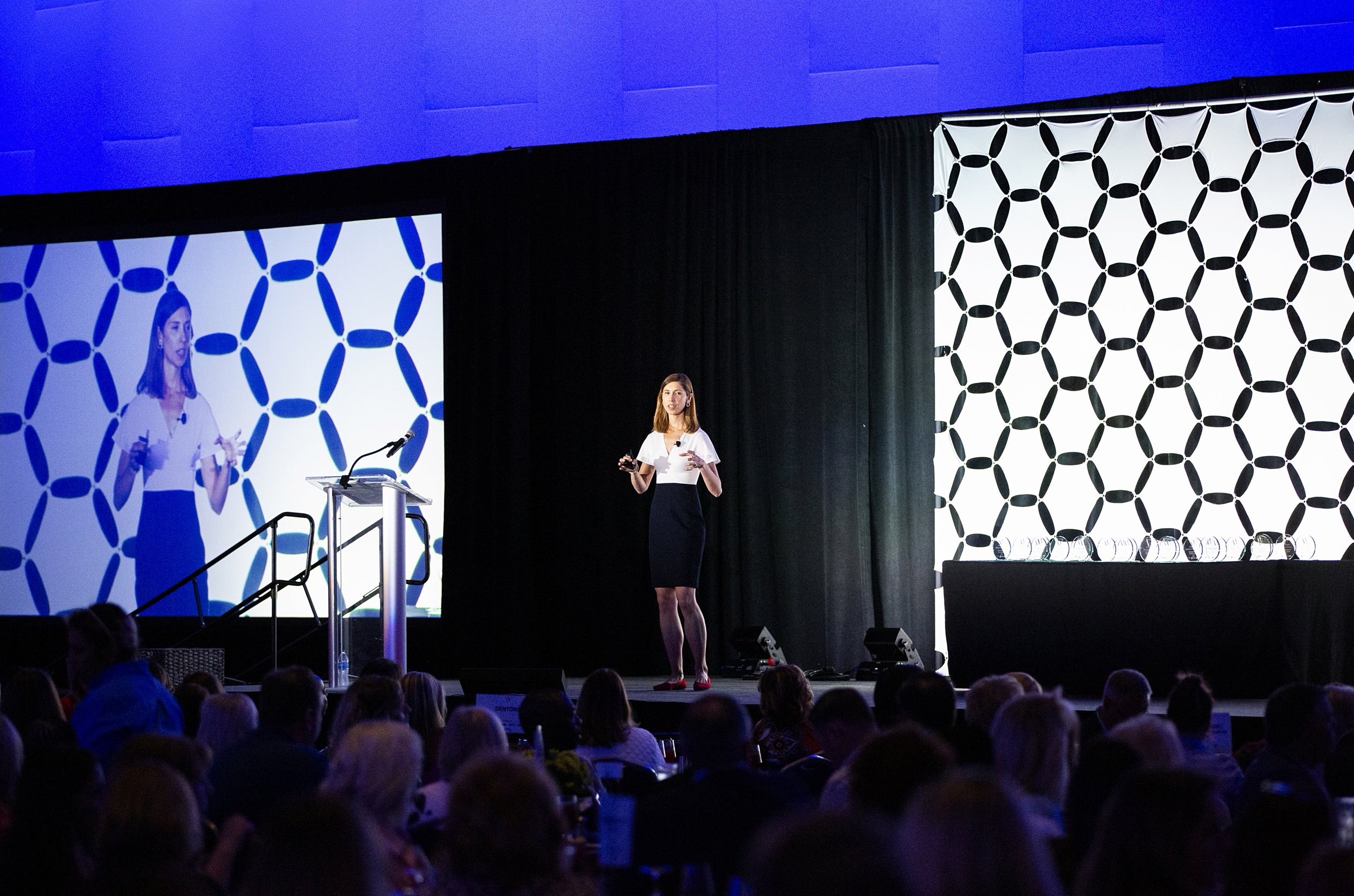 Kristel Bauer giving a presentation on stage at a conference with a large screen behind her displaying a patterned black and white background. Audience members are seated in front.