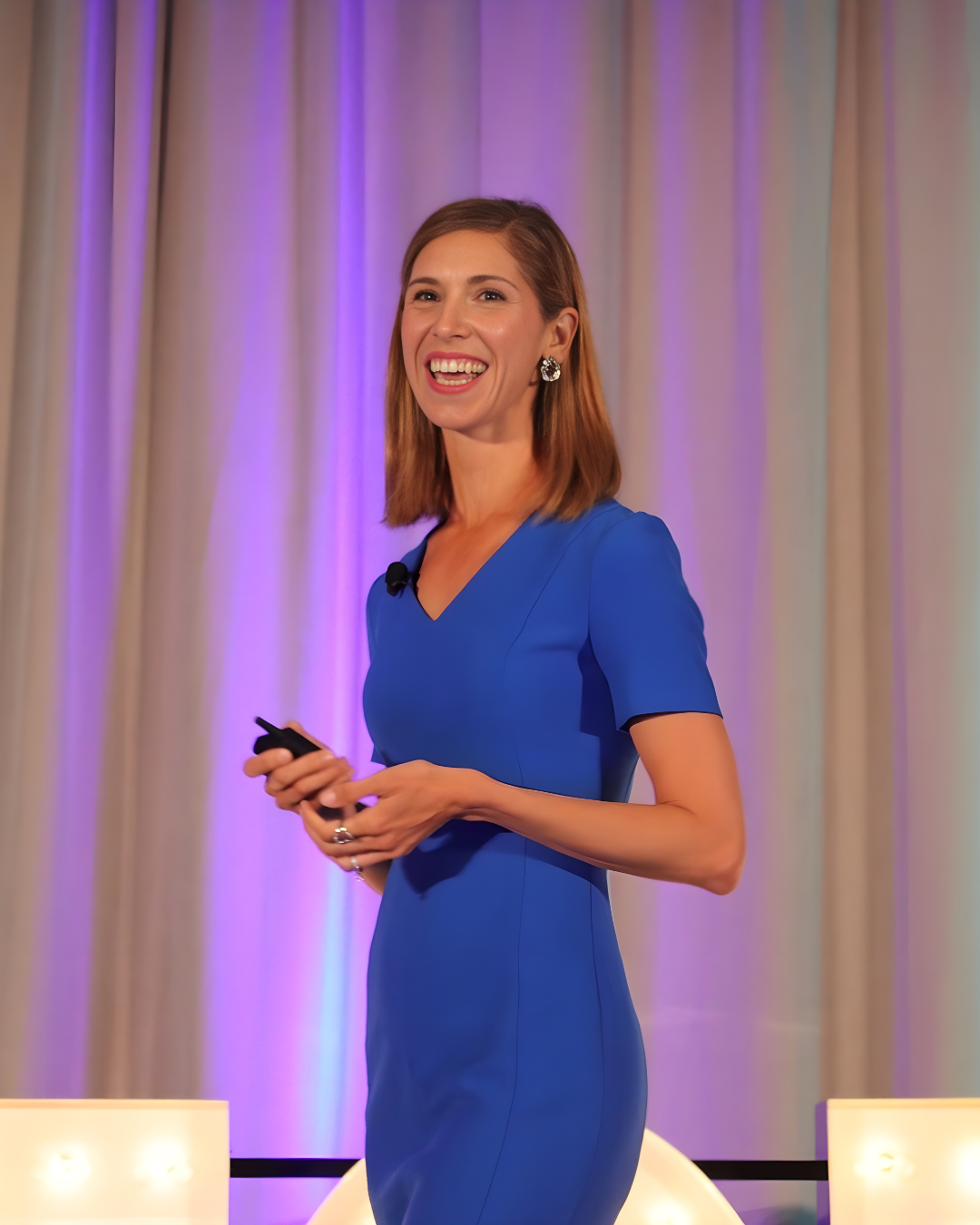 Kristel Bauer, a woman in a blue dress smiling and holding a microphone on stage in front of curtains with purple lighting.