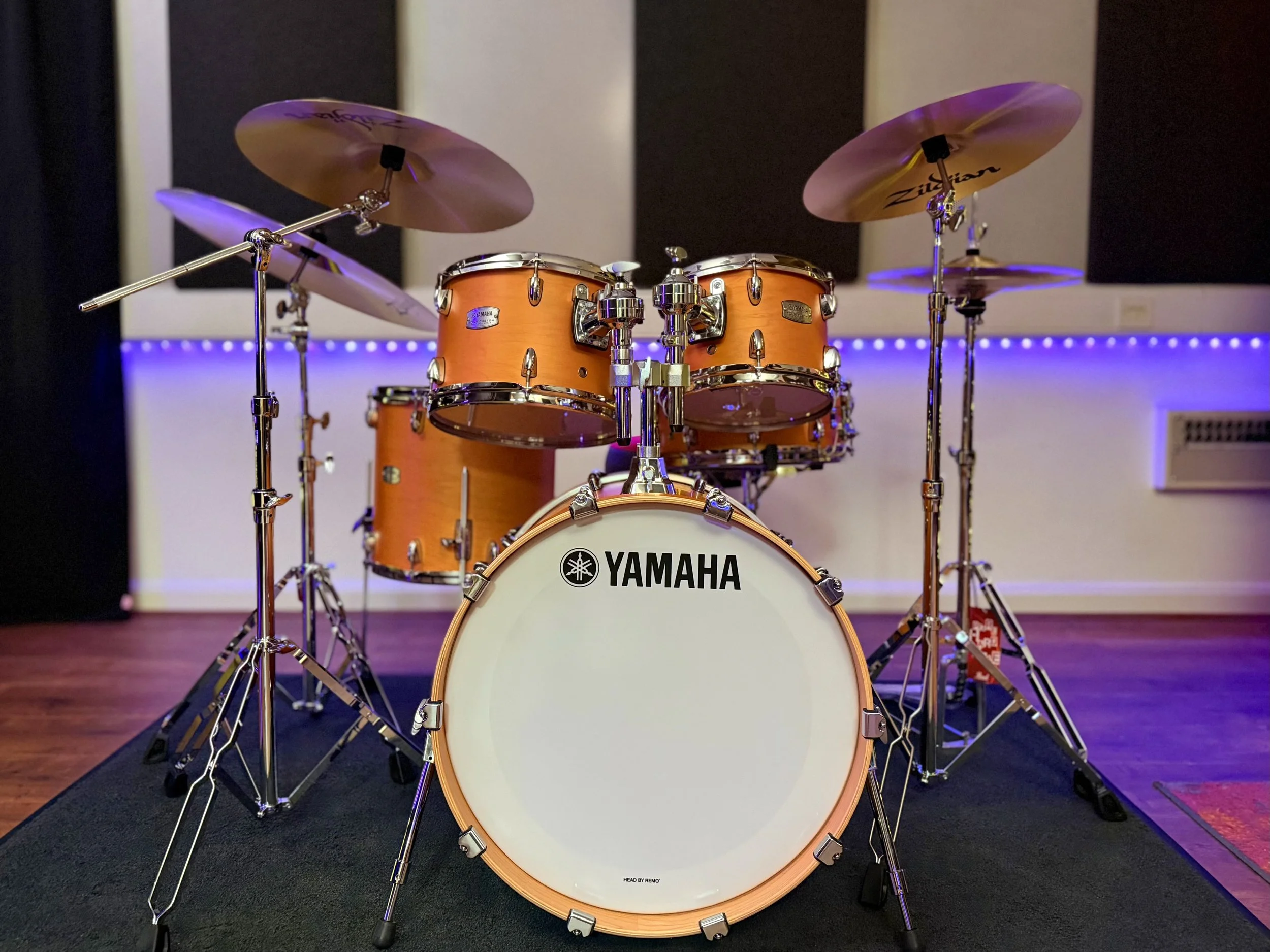 An orange Yamaha drum set with cymbals on stands, positioned on a black mat in a room with black and white walls and purple lighting accents.