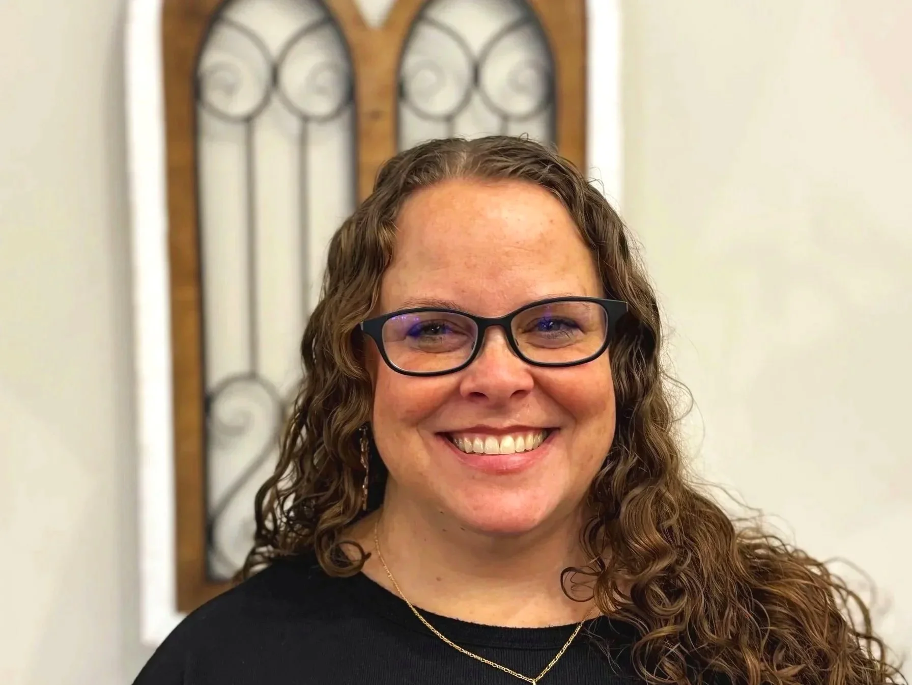 A woman with curly brown hair, wearing glasses, earrings, a gold necklace, and a black top, smiling indoors with a decorative wrought iron and wood piece behind her.