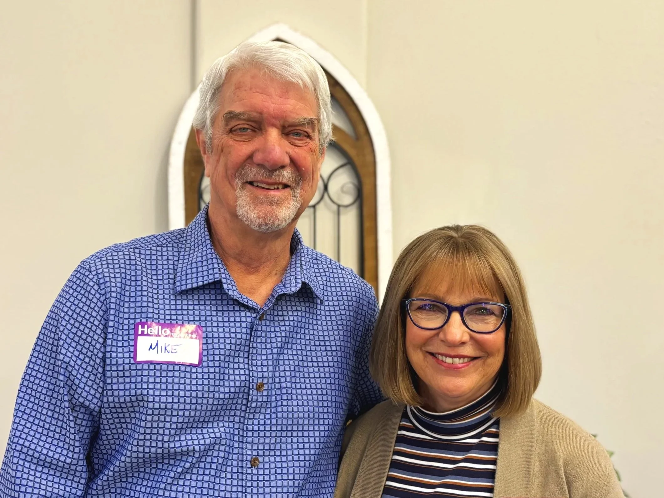 A smiling middle-aged man with gray hair and beard, wearing a blue checkered shirt with a name tag that says 'Mike', standing next to a smiling woman with blonde hair, glasses, a striped turtleneck, and a beige blazer, in an indoor setting with a decorative window behind them.
