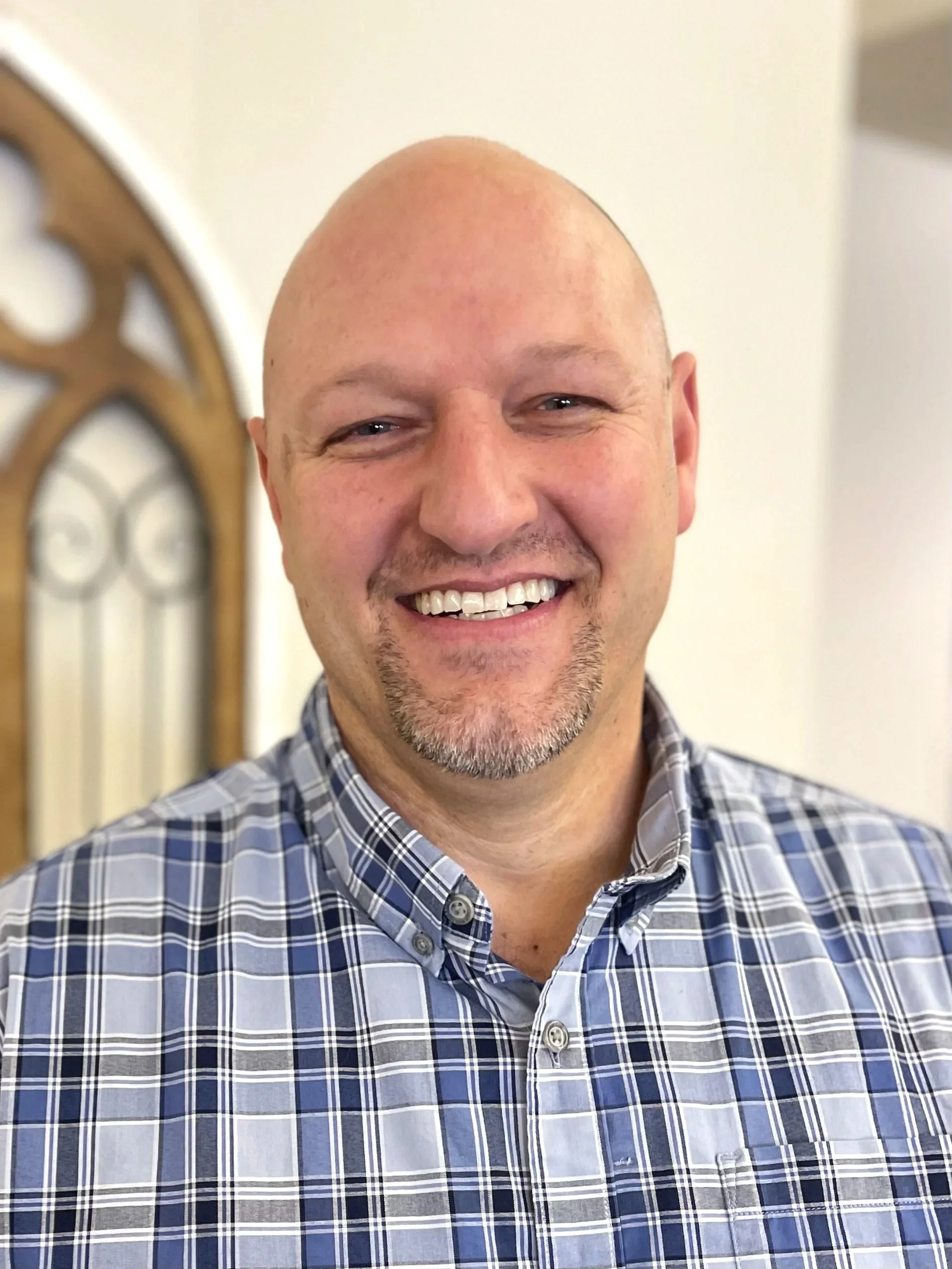 A smiling man with light skin, a bald head, and a goatee, wearing a plaid shirt, standing indoors in front of a decorative mirror or window.