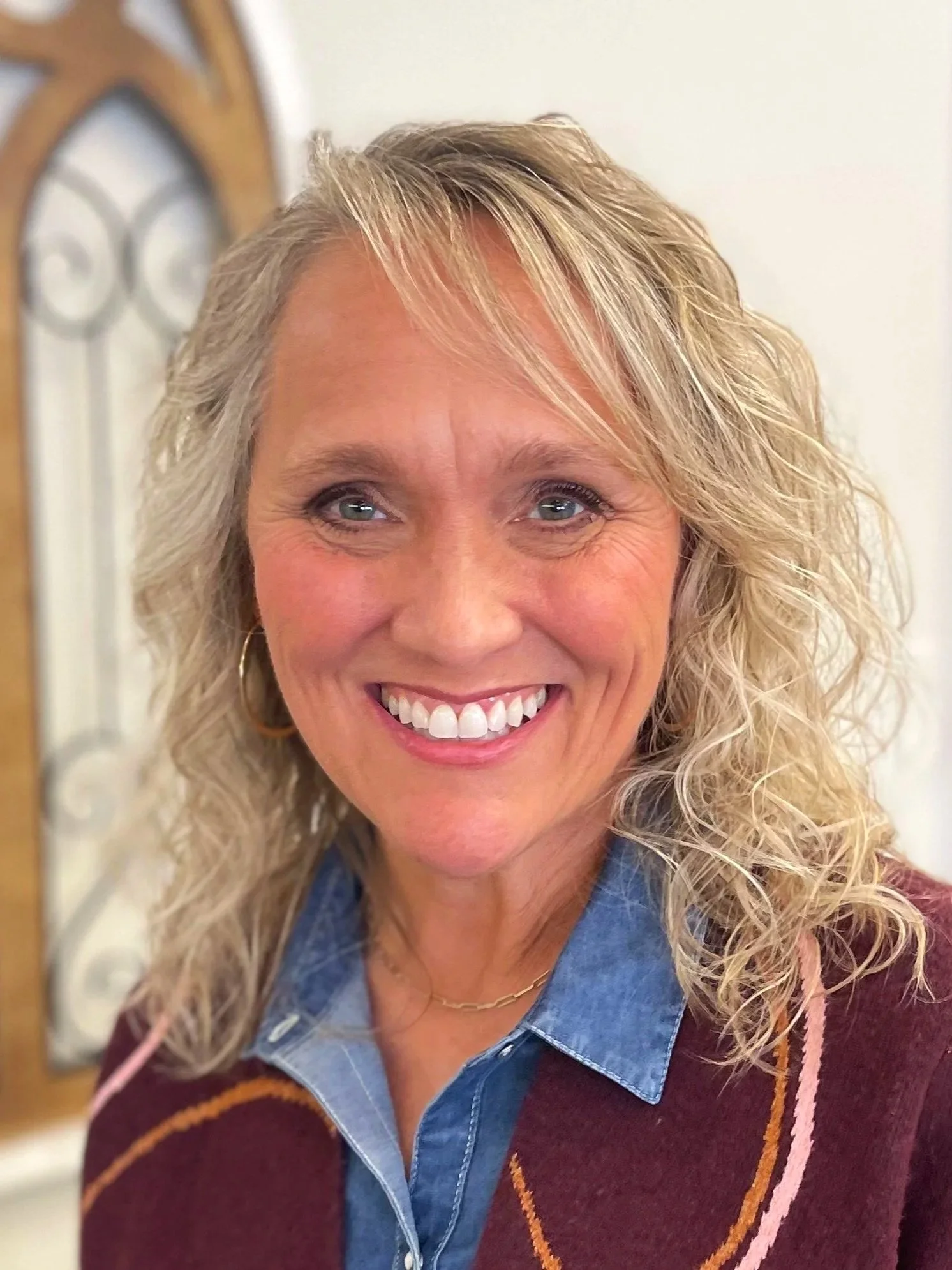 A smiling middle-aged woman with curly blonde hair, wearing a denim shirt and a maroon sweater with orange and pink accents.