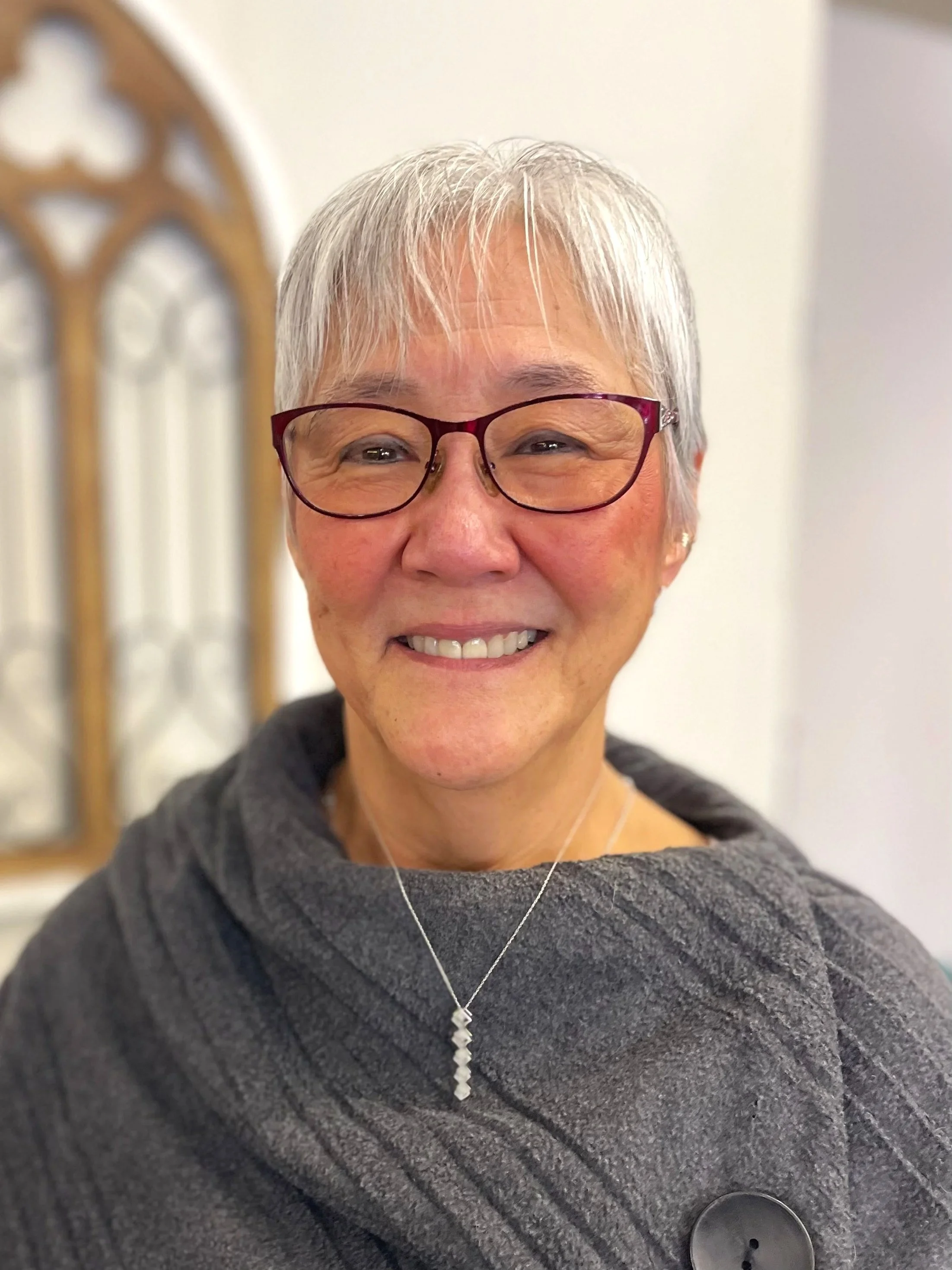 A smiling older woman with short gray hair, wearing glasses, a gray shawl or sweater, a silver necklace with a pendant, and earrings.