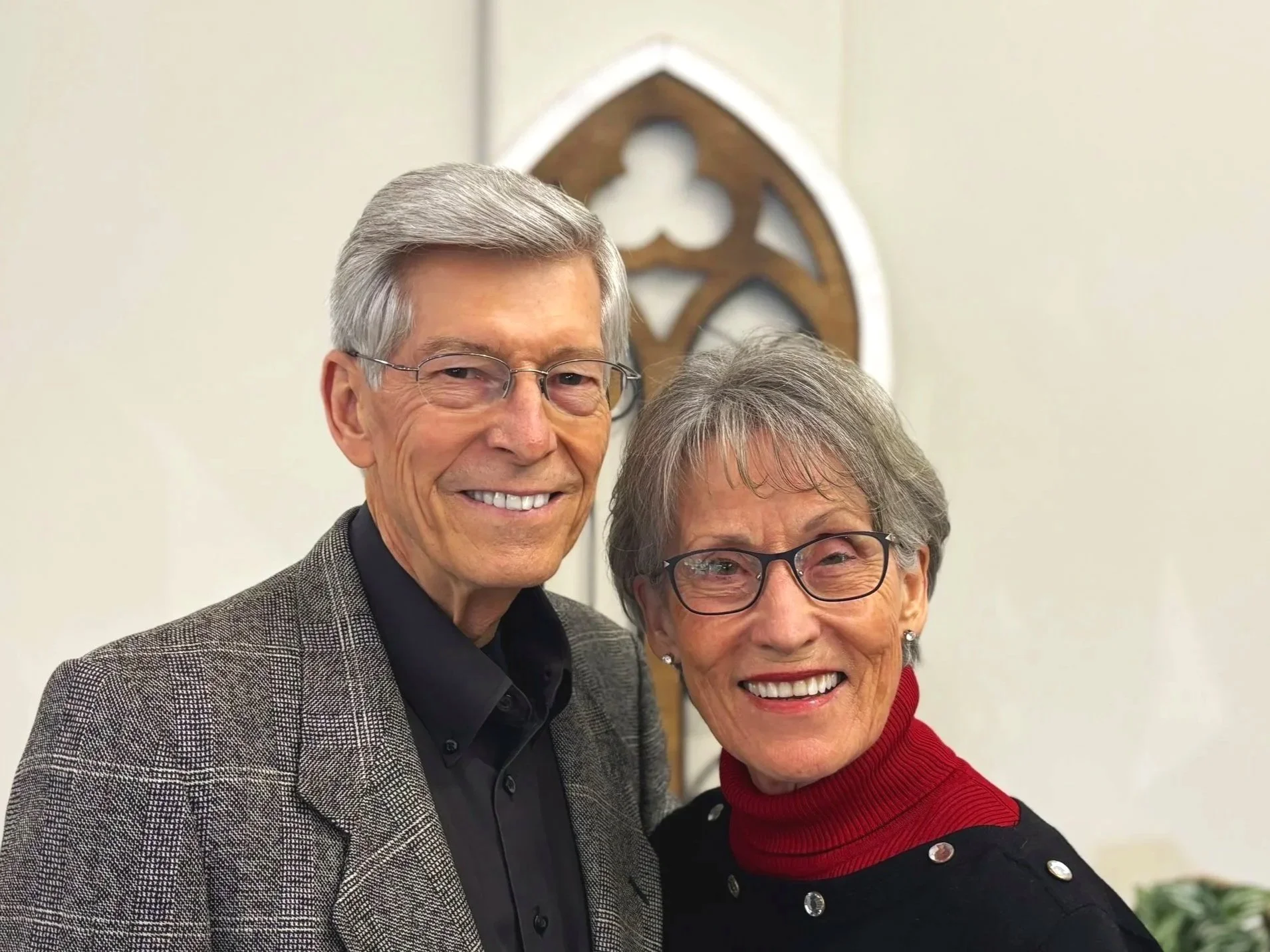 Smiling elderly man with gray hair and glasses standing next to smiling elderly woman with gray hair and glasses, both in front of a church interior with a wooden decorative element on the wall.