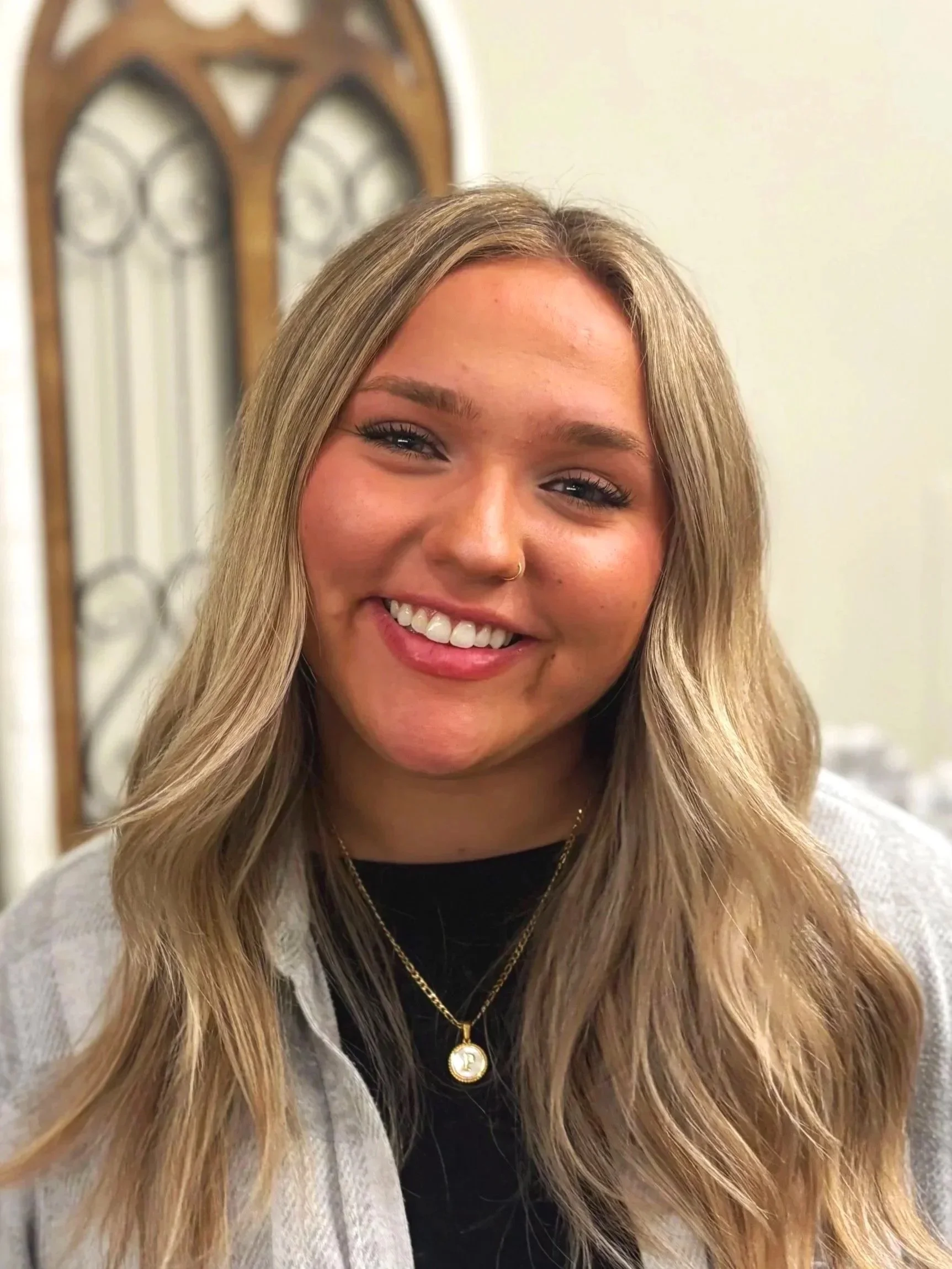 Close-up of a young woman with long wavy blonde hair, smiling, wearing a black top, a light-colored jacket, a gold necklace, and a nose ring. Background shows a decorative mirror with a wooden frame.