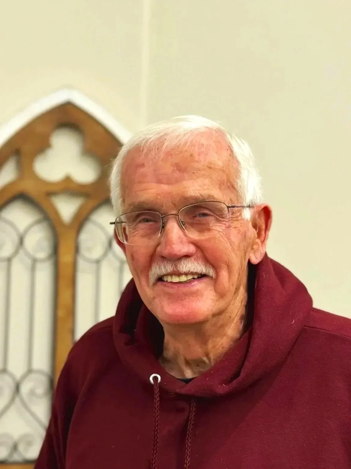 Smiling elderly man with gray hair, glasses, and a mustache, wearing a maroon hoodie in front of a stained glass window.