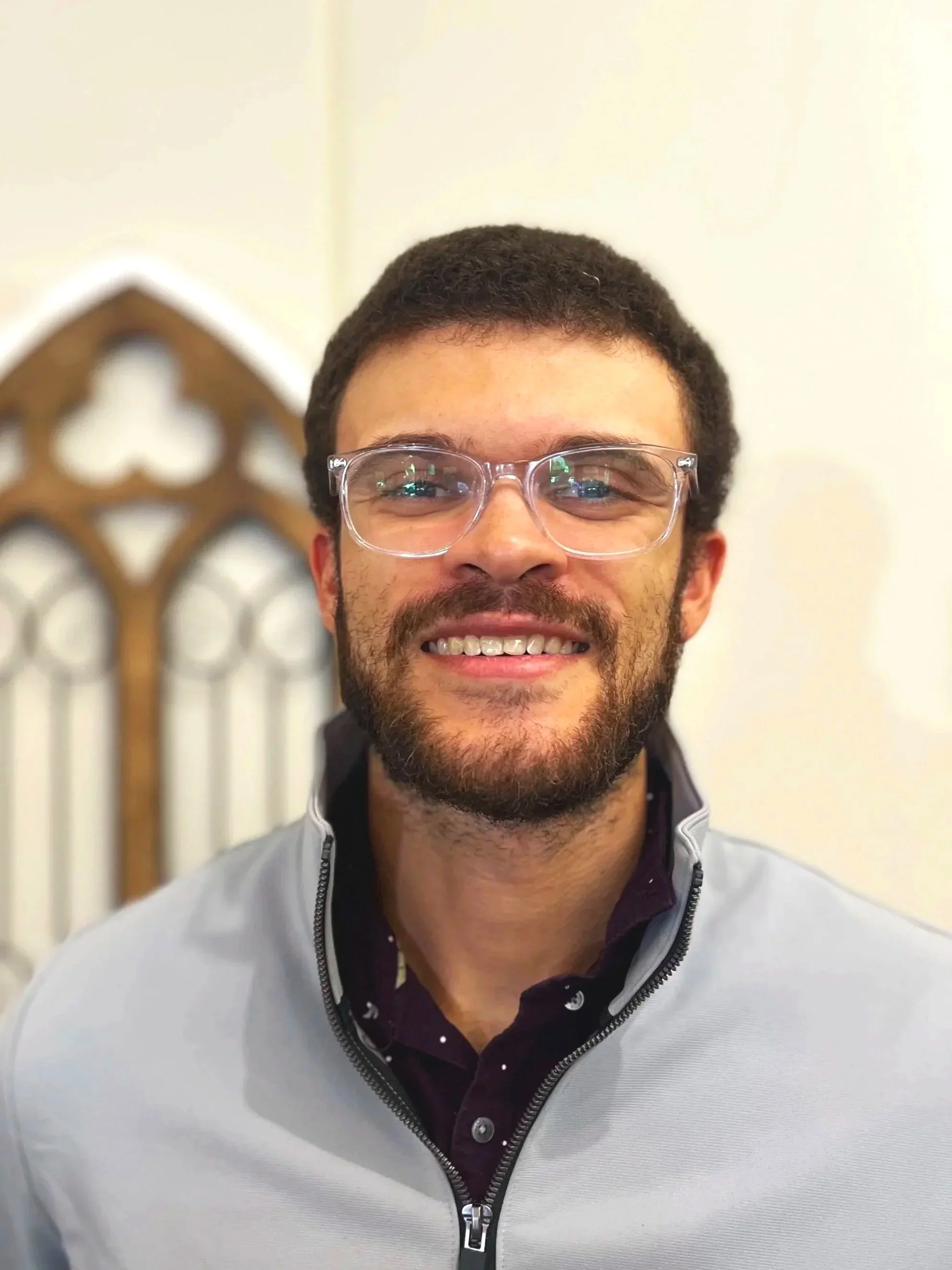 A smiling man with glasses, a beard, and short dark hair, wearing a light gray jacket over a dark shirt with a pattern, standing in front of a decorative wooden headboard or architectural detail.