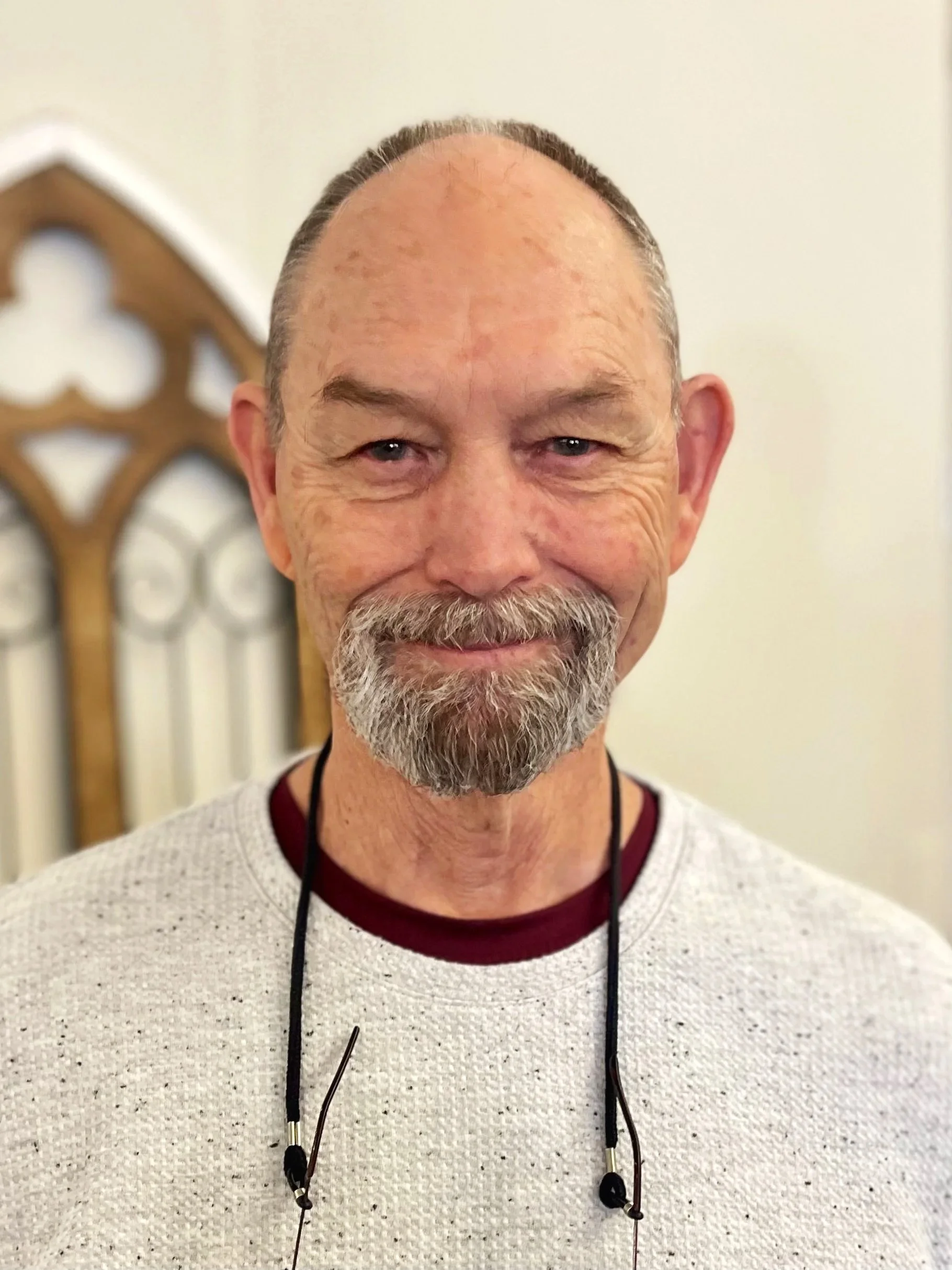 A smiling older man with a beard and mustache wearing a white sweater and glasses hanging from a lanyard around his neck, standing indoors.