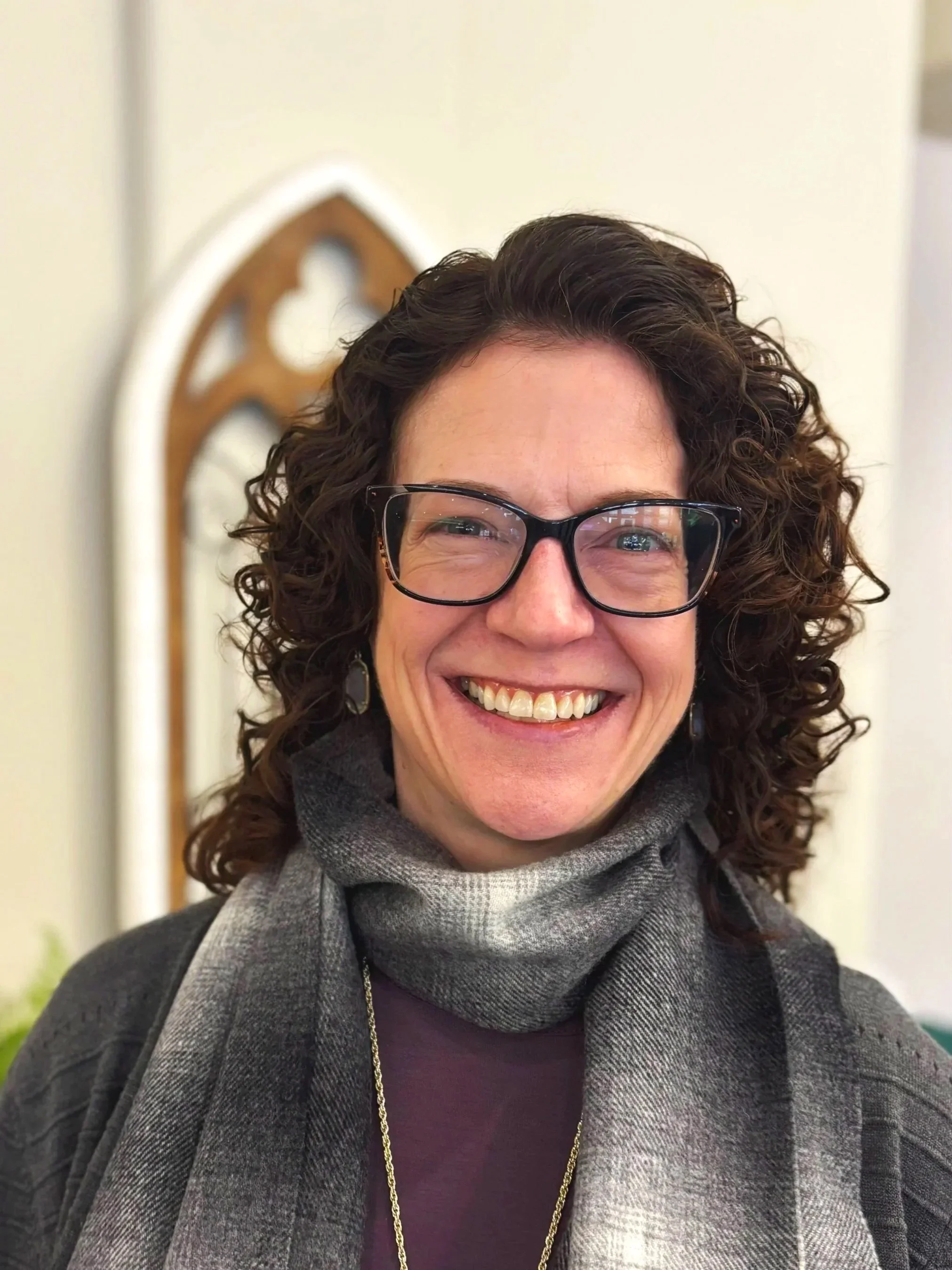 A smiling woman with curly dark hair and glasses. She is wearing a gray scarf, a dark top, and a gold chain necklace, standing indoors with a background featuring a decorative white and brown mirror.