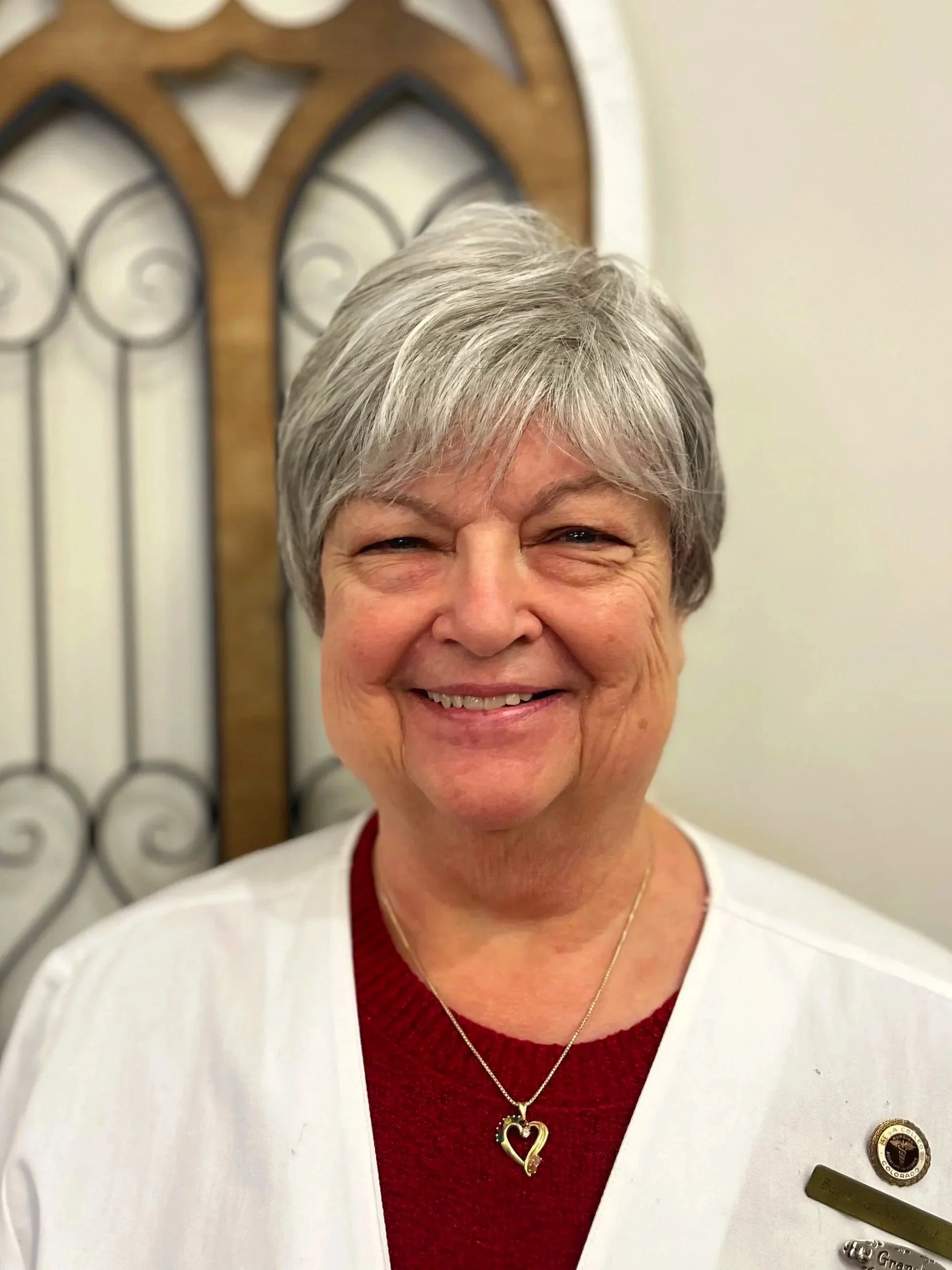 A smiling older woman with short gray hair, wearing a white coat, a red shirt, a gold heart-shaped necklace, and a name badge, standing in front of a decorative wooden and metal headboard.
