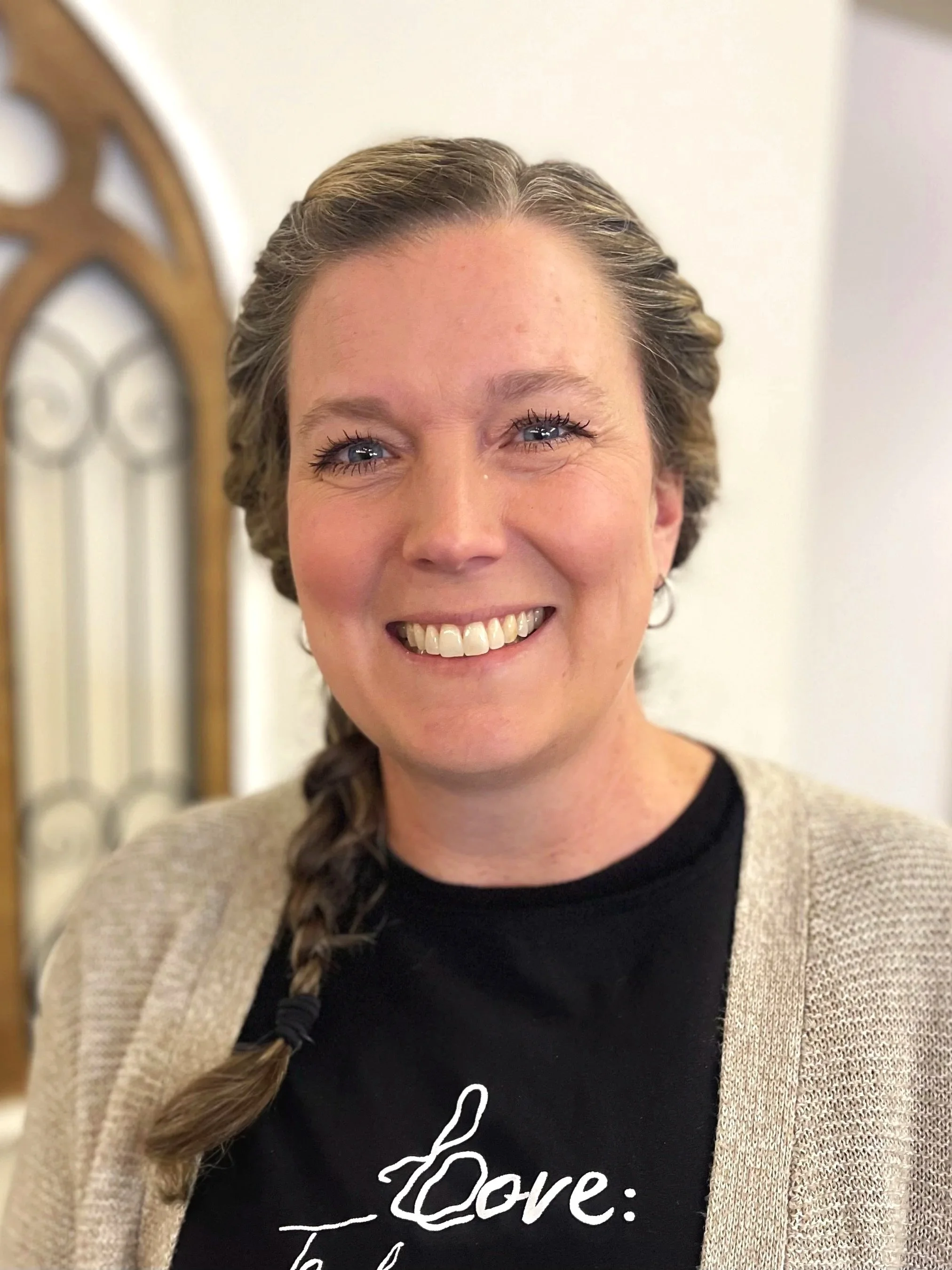 A woman with blue eyes and a braid, smiling, wearing a black shirt with white writing and a beige cardigan, standing in front of a mirror or decorative wooden frame in an indoor setting.