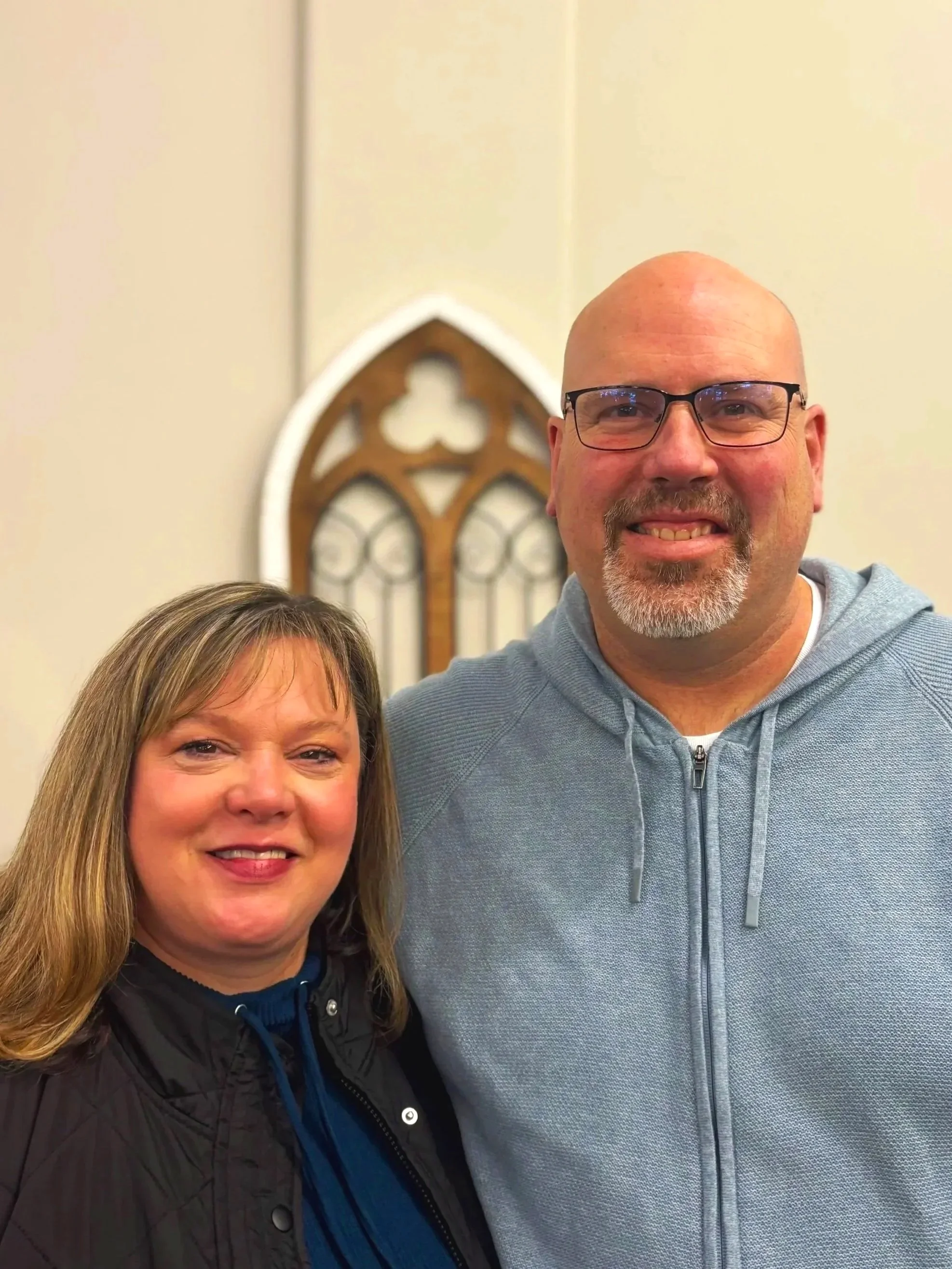 A smiling woman and man standing together indoors in front of a church window. The woman has shoulder-length blonde hair and is wearing a black jacket with a blue top. The man is bald, wearing glasses, and a light gray zip-up hoodie.