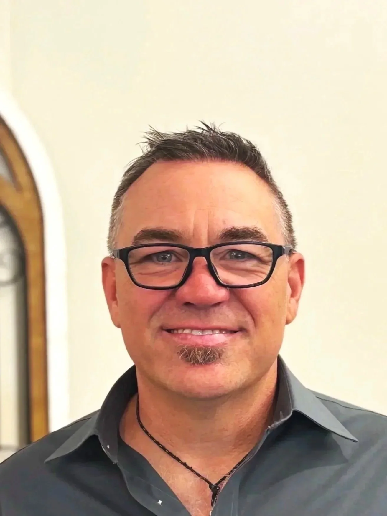 A man with short dark hair, glasses, and a goatee, smiling in front of a plain light-colored wall.