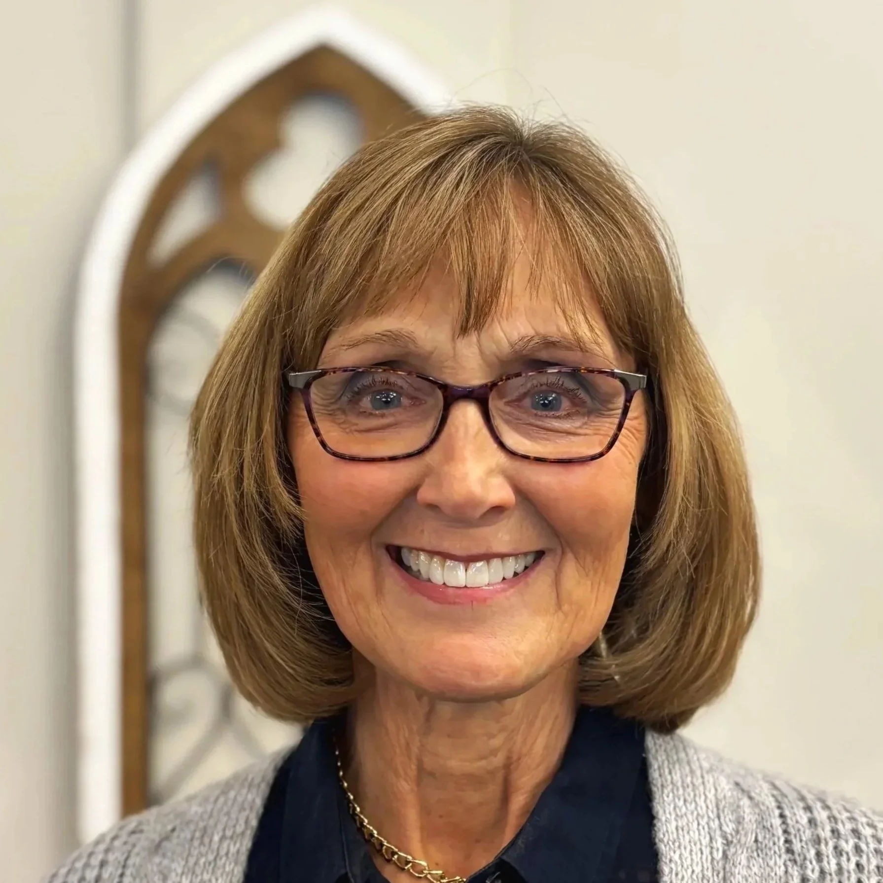 A smiling elderly woman with glasses and short, reddish-brown hair. She is wearing a gray sweater over a dark-colored shirt and a gold necklace. There is a wooden decorative piece in the background.