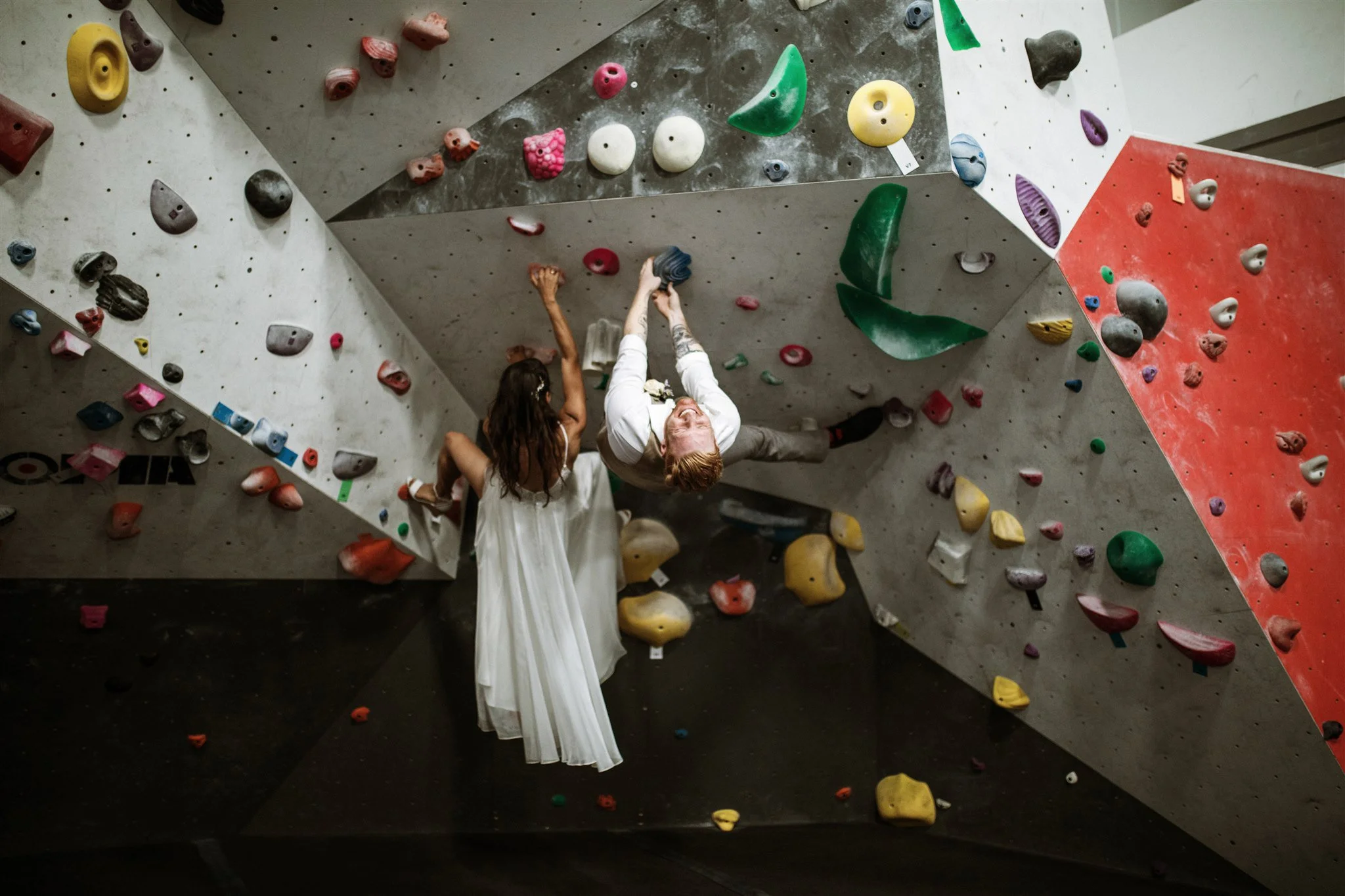 Two people, one woman in a white dress and another man in a white shirt and beige pants, are rock climbing indoors on a structured bouldering wall with colorful holds.