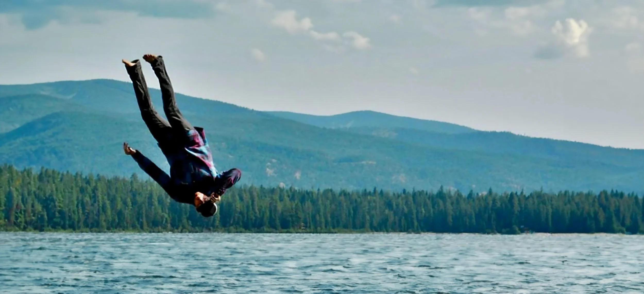 Person performing a backflip jump over a body of water with forest and mountains in the background.