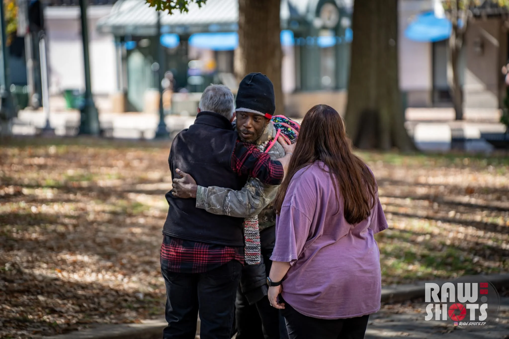 A man in a black beanie and camouflage jacket hugging an elderly man in a black vest, while a woman with long brown hair and a purple shirt stands nearby. They are outdoors in a park with trees and fallen leaves.