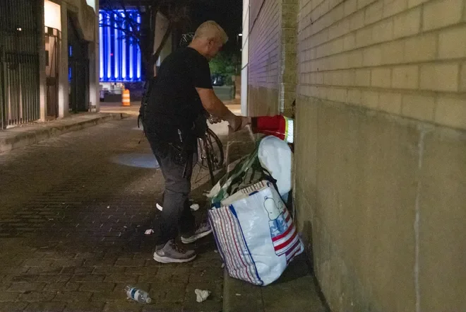 A man cleaning a red fire hydrant on a city sidewalk at night, with a large bag and a plastic bottle on the ground nearby.