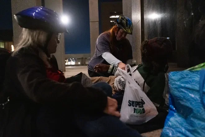 Three people sitting on the floor in an indoor setting, wearing helmets, with bags and food bags around them.