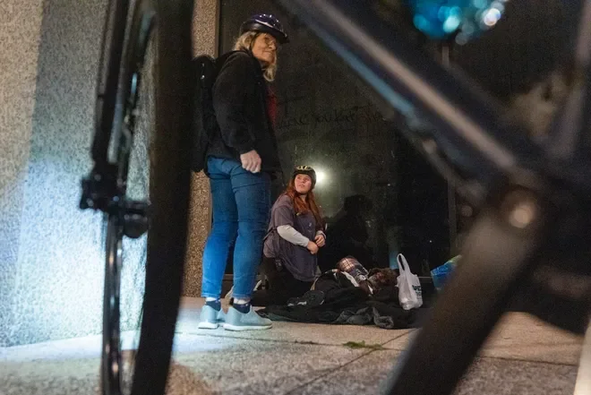 Two women wearing helmets are sitting and standing on a sidewalk at night near an urban building, with personal belongings and bags on the ground.