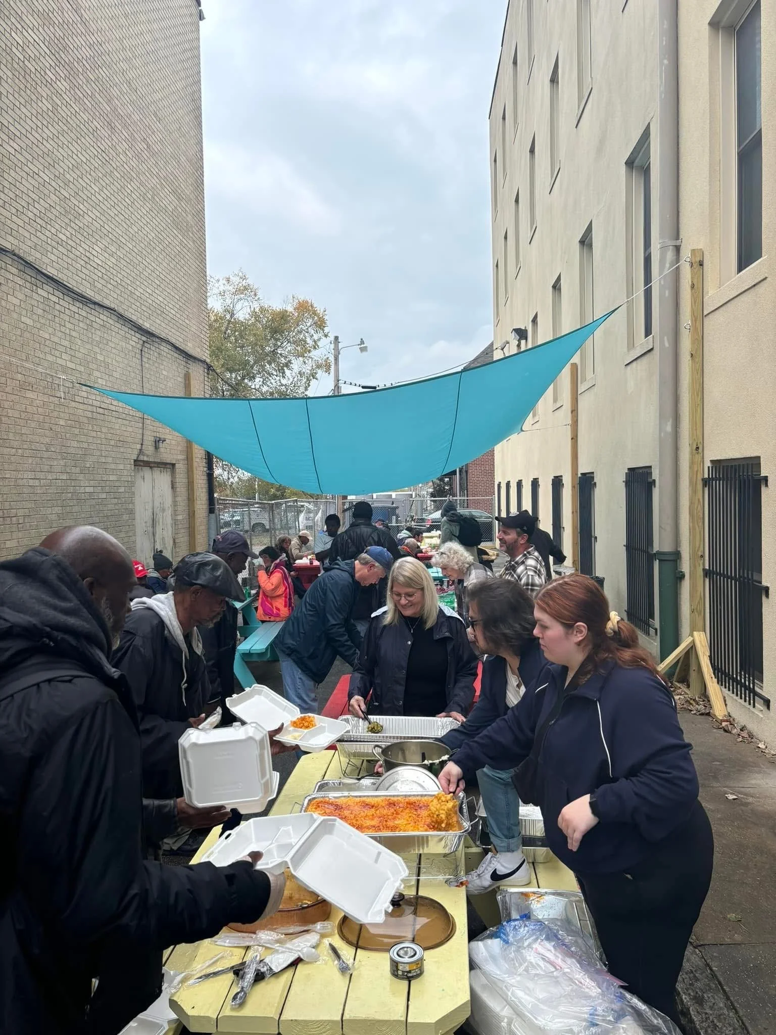 People gathered in an alleyway under a large blue canopy, serving food from trays at a community meal or event.
