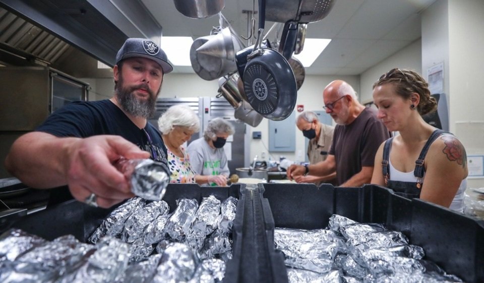 People in a kitchen preparing food, wrapping items in foil, with oven mitts and cooking utensils hanging above.