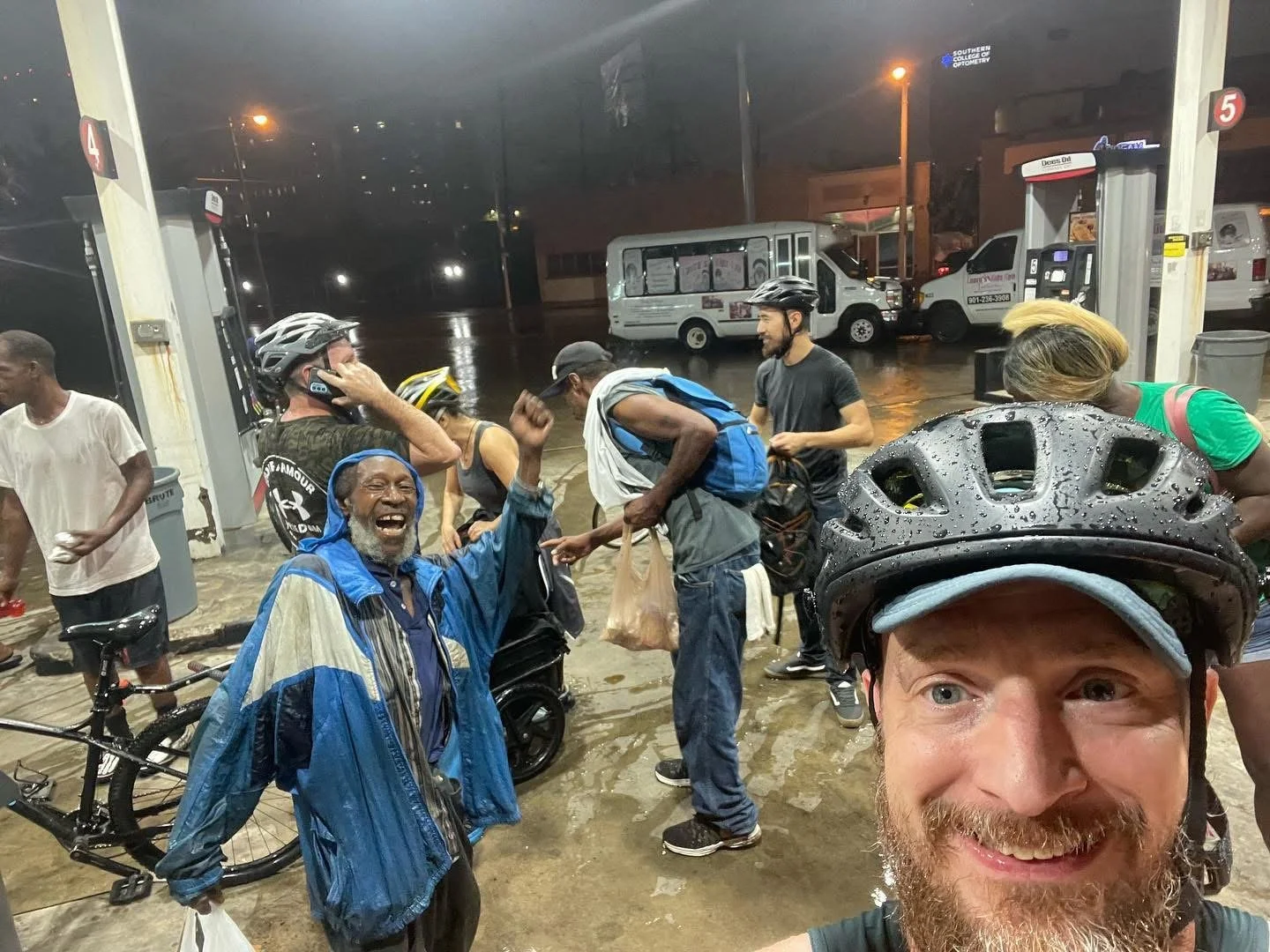 Group of cyclists and pedestrians under a rain-soaked gas station canopy at night, some wearing helmets and rain jackets, with a man in the foreground smiling and taking a selfie, and an older man laughing with raised arm. Wet ground and vehicles in 