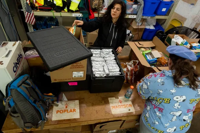 A woman at a checkout counter packing food items wrapped in aluminum foil, with a young child dressed as a police officer standing nearby. The scene appears to be in a storage or donation center.
