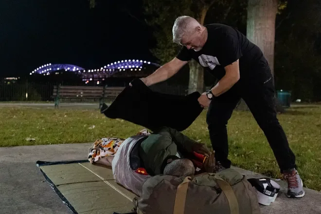 A man in black clothing is helping a person lying on the sidewalk, covered with a blanket, in a park at night. There are trees, a bench, and city lights in the background.