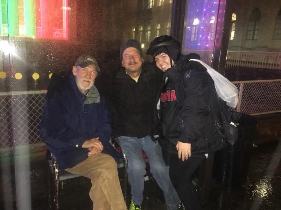 Three people sitting on a bench outdoors at night in the rain, smiling, with a glass building and colorful lights in the background.