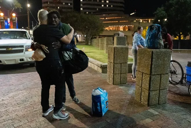 Two people hugging on a sidewalk at night, with others in the background and a large backpack on a ledge.