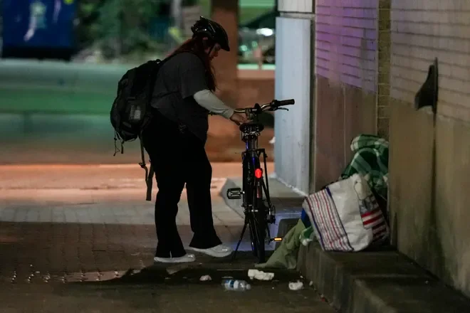 A person wearing a helmet, black backpack, and black clothing, standing on a sidewalk and adjusting a bicycle next to a wall at night.