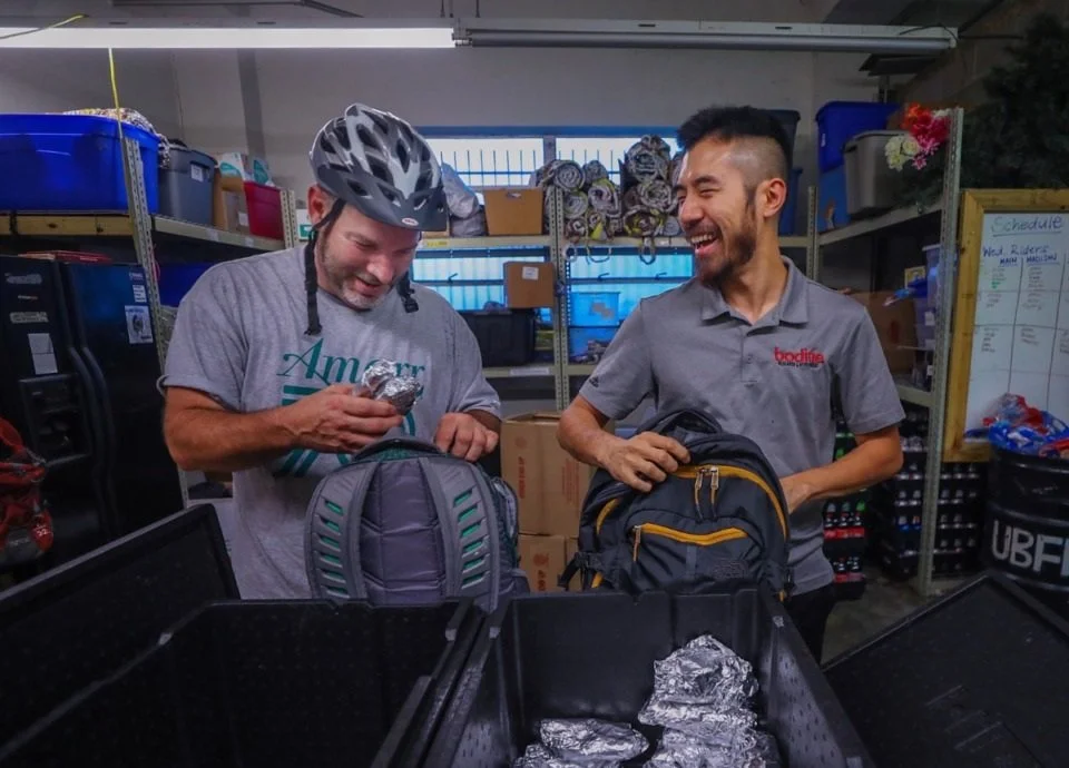 Two men in a storage room are smiling and sharing a moment while holding backpacks and eating foil-wrapped food; one is wearing a bicycle helmet, and shelves with boxes and supplies are visible in the background.