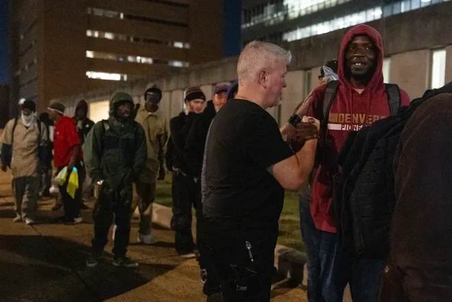 A group of people, mostly men, standing in line outdoors at night, while a man in a black shirt interacts with a young man in a red hoodie.