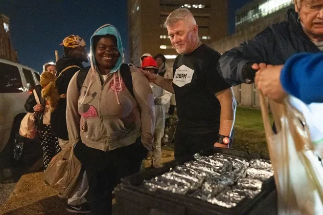 People gathered outdoors at night, with one smiling woman wearing a hoodie and backpack in the foreground, and a man next to her wearing a black shirt and a watch, standing behind a table with silver-colored packages. Several other people are visible