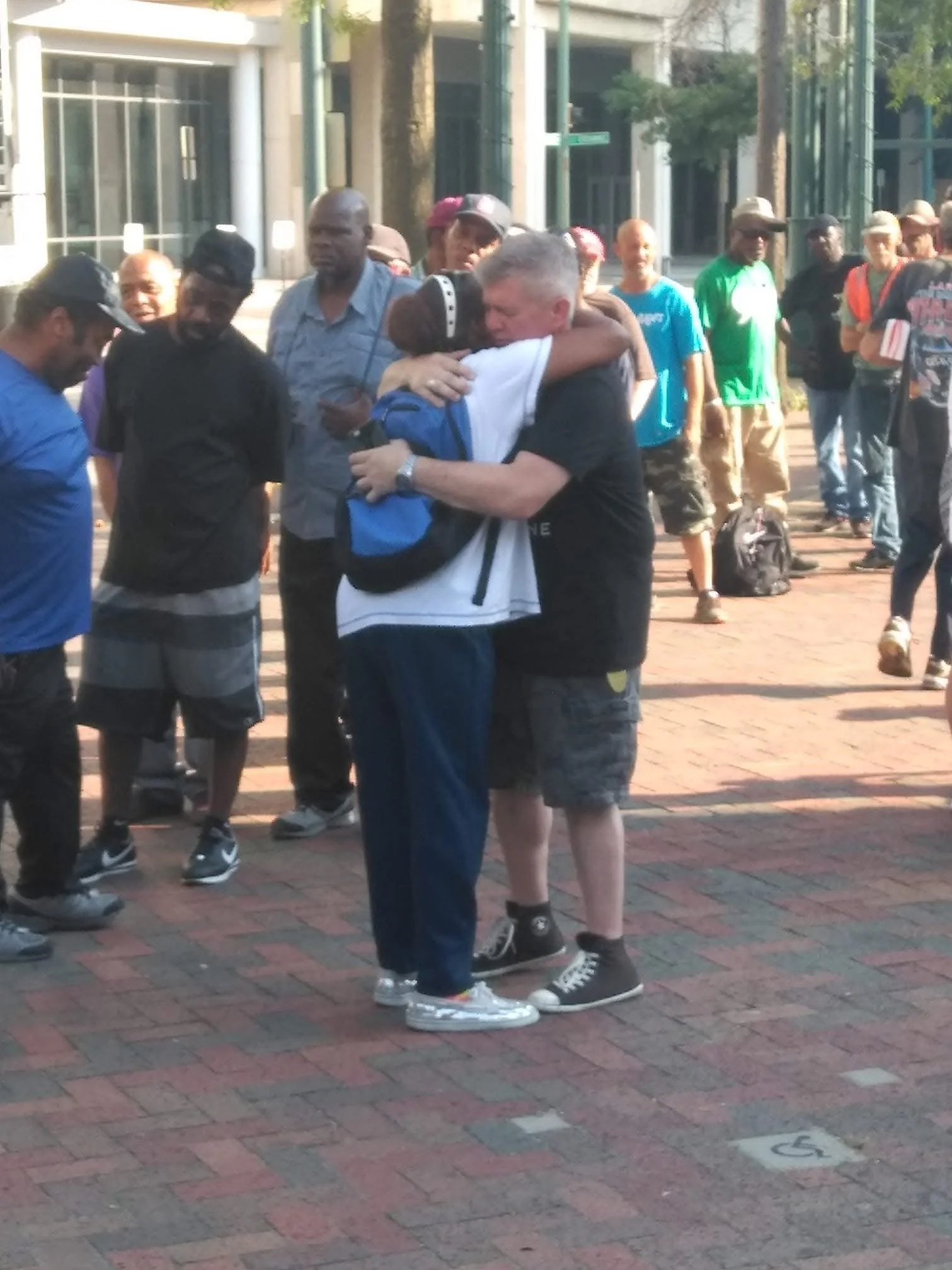 Two people hugging in the center of a group of diverse onlookers on a brick sidewalk in an urban area.
