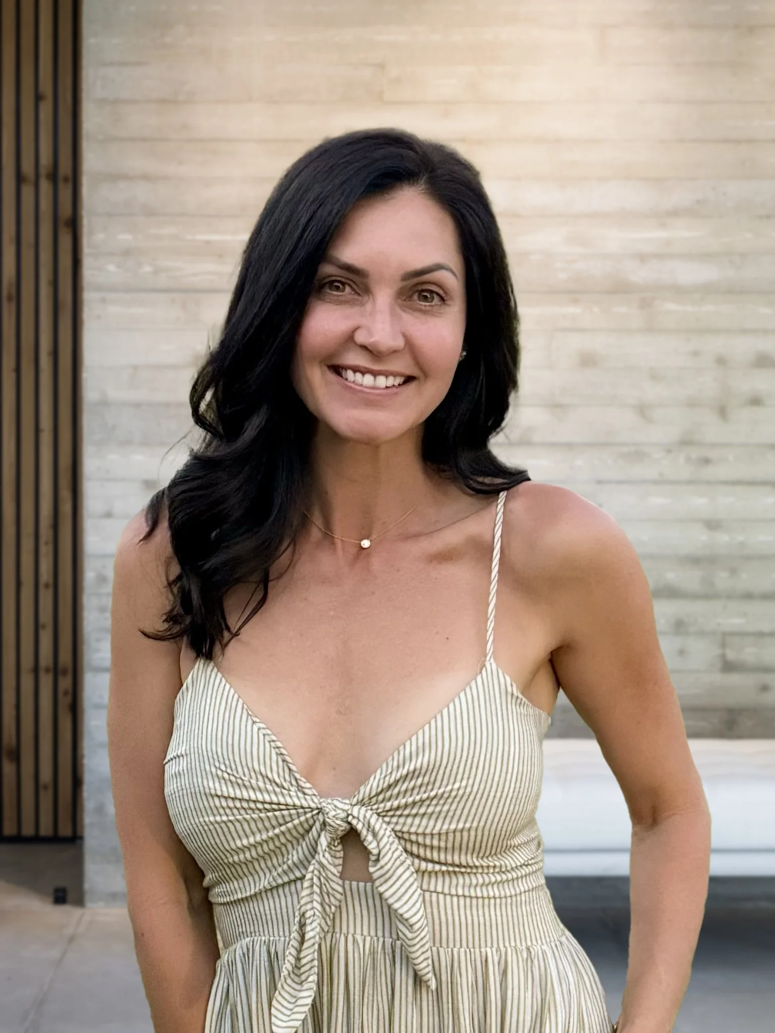 A woman with black hair smiling, wearing a striped dress with spaghetti straps and a bow, standing outdoors in front of a modern building with wooden paneling.