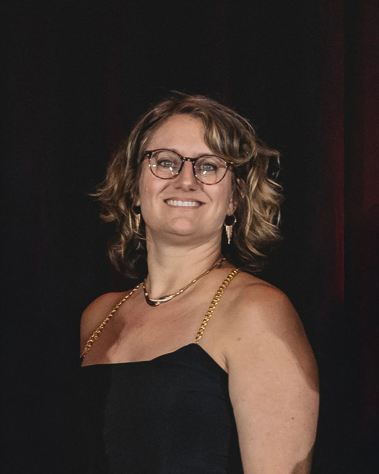 Woman with wavy light brown hair, glasses, gold jewelry, and a black dress, smiling on a dark background.