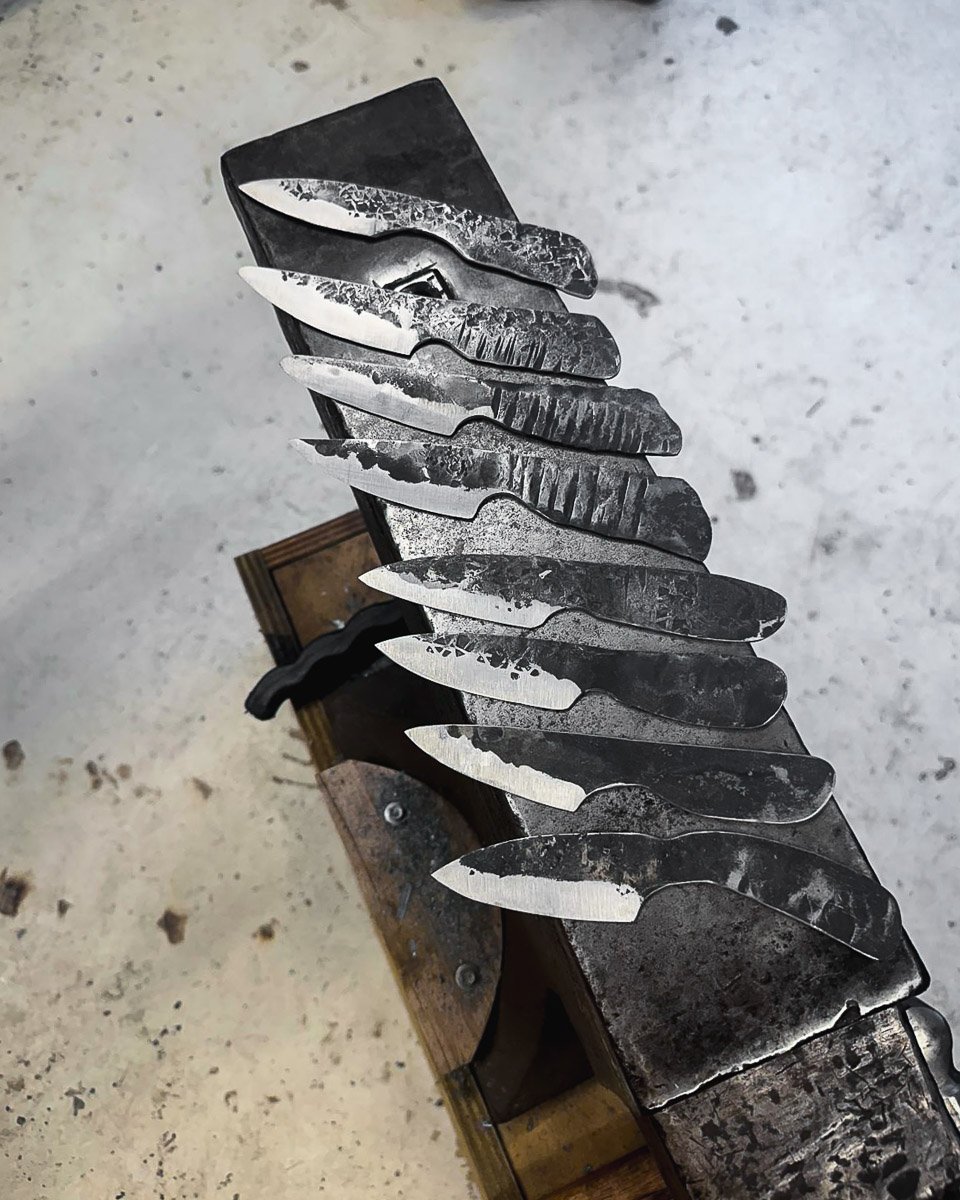 A collection of rustic, forged knives with various blade shapes and textures, resting on an anvil.