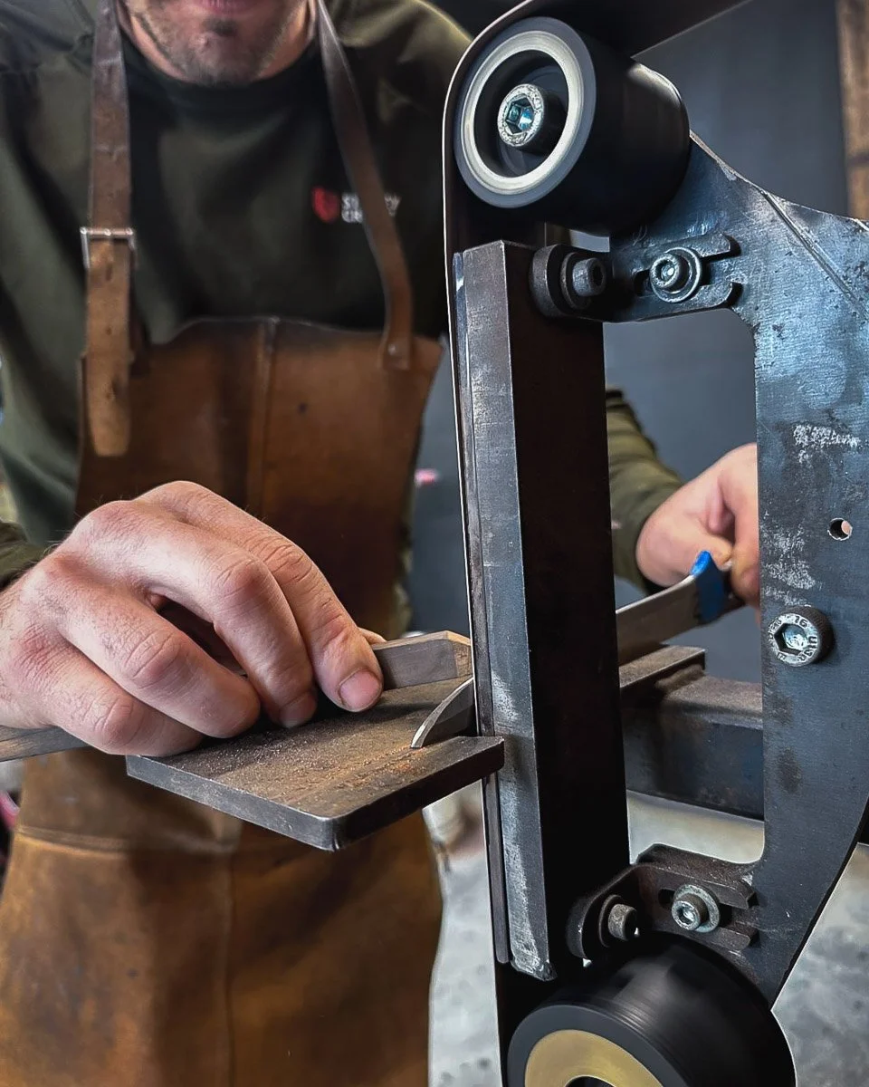 A person is sharpening a small knife on a belt grinder machine in a workshop.