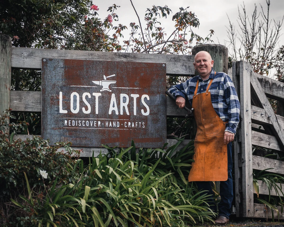 A man standing outdoors next to a rustic wooden fence and a metal sign that reads "Lost Arts Rediscover Hand-Crafts." The man is wearing a plaid shirt, brown apron, and jeans, and is smiling while holding a tool with his right hand.