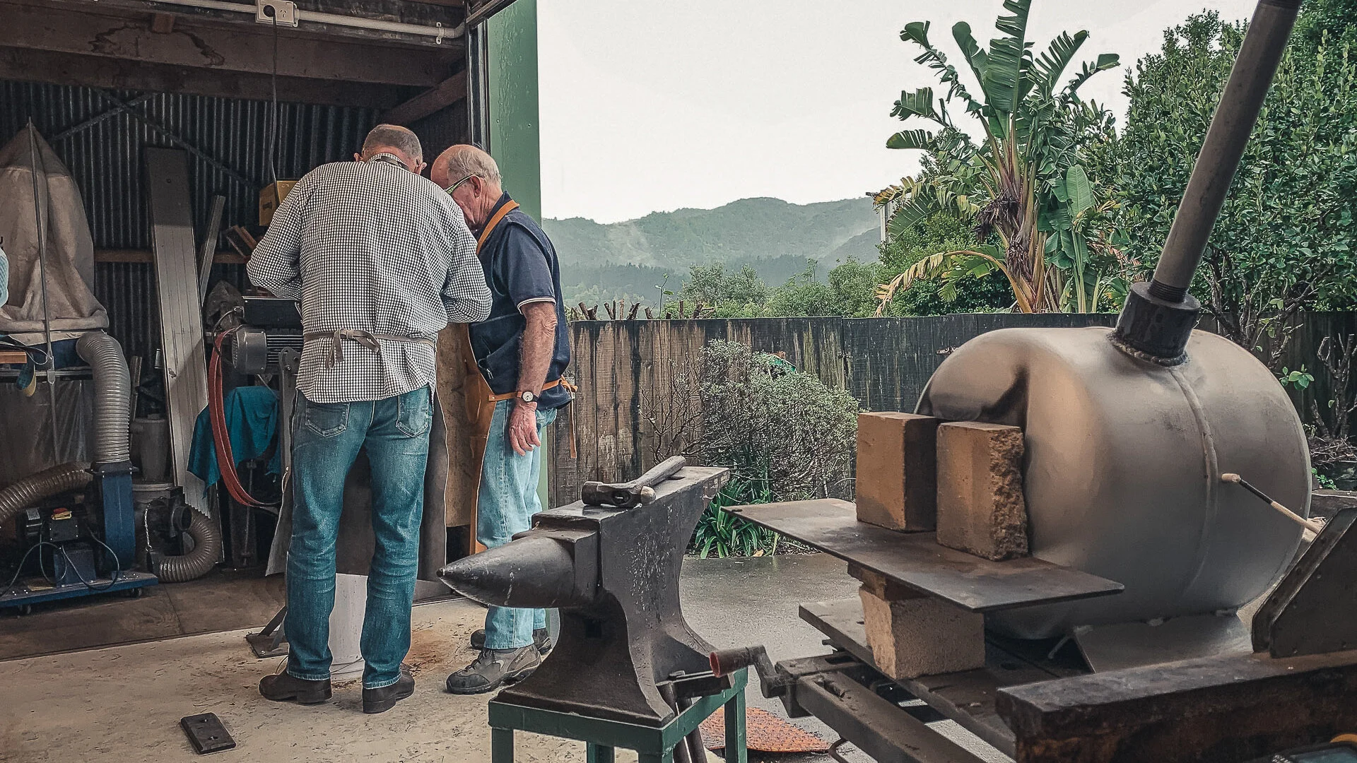 Two men are working together in a workshop, focused on a task. One man wears glasses and a checkered shirt, while the other has gray hair and a beard, wearing a dark shirt and apron. The workshop has a mountain landscape visible in the background, with green trees and a banana plant outside. In the foreground, there is a metal drum connected to a pipe, and an anvil with a hammer on top.