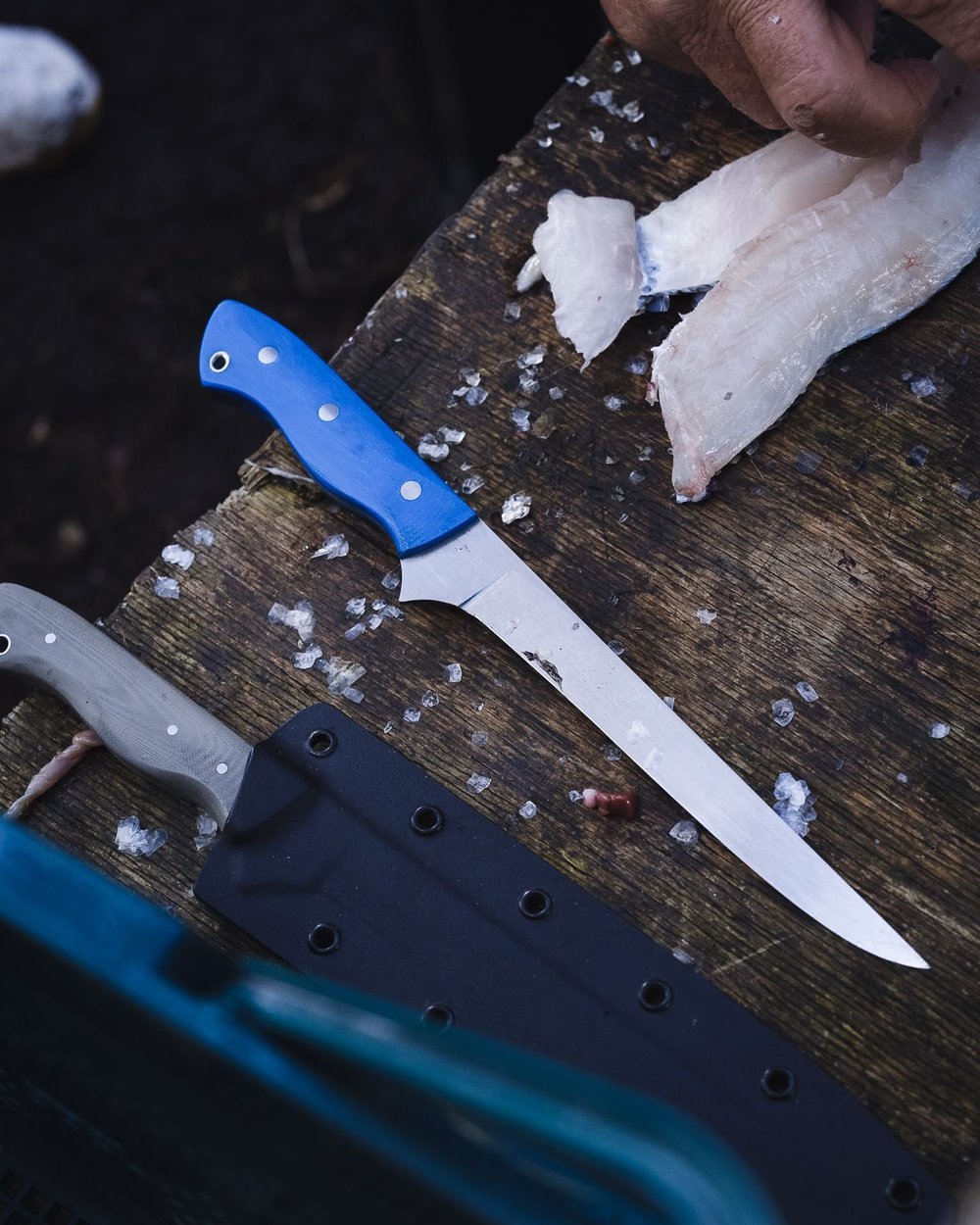 Handmade stainless filleting knife with a blue micarta handle beside its black Kydex sheath, on a board with fish fillets