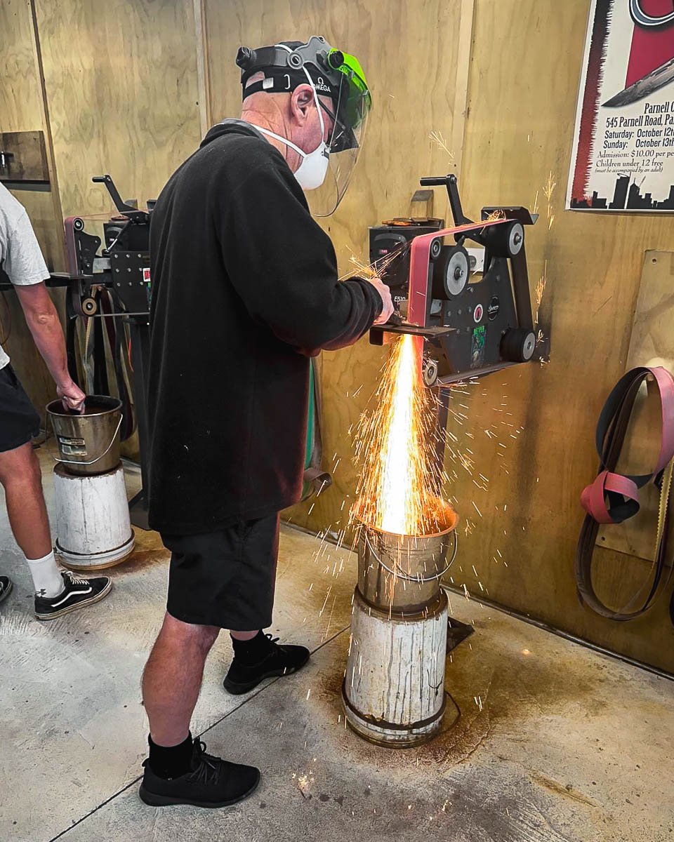 Man grinding a knife with sparks flying, wearing safety helmet and face mask, in workshop setting.