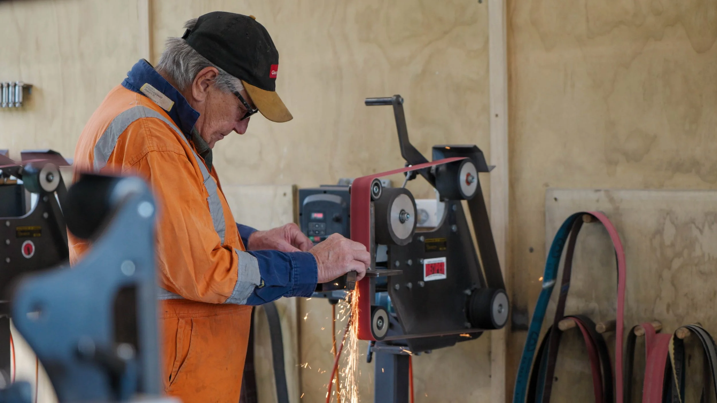 An elderly man wearing safety glasses, a black cap, and orange work overalls grinding a piece of metal, with sparks flying in a workshop setting.