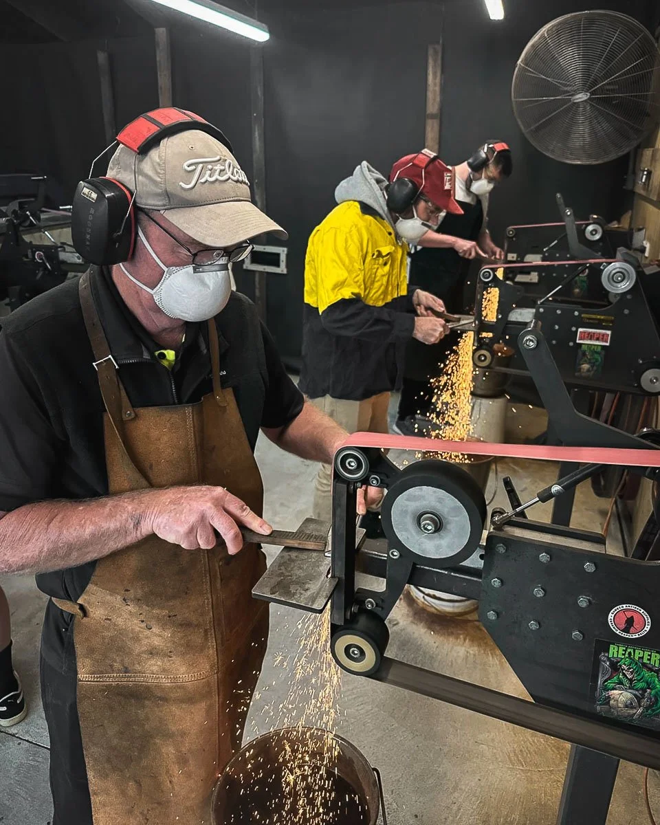 People working with a machine that cuts or shapes a material, with sparks flying down. They are wearing face masks, ear protection, and gloves, in a workshop or factory environment.